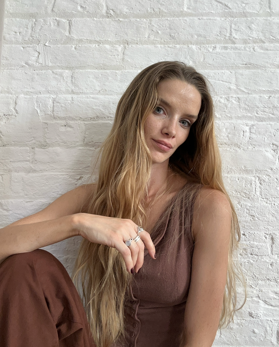 A woman with long wavy blonde hair sitting against a white brick wall, wearing a sleeveless brown top and multiple silver rings, looking at the camera.