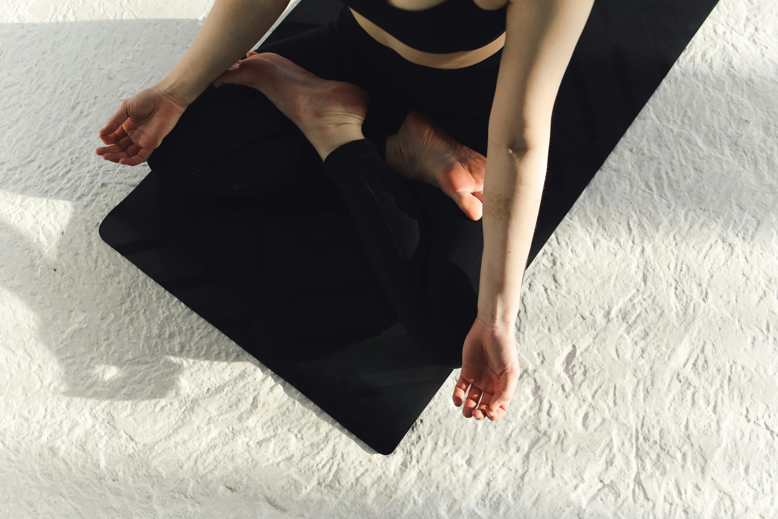 Person practicing yoga in a cross-legged position on a black mat, with a textured white floor underneath.