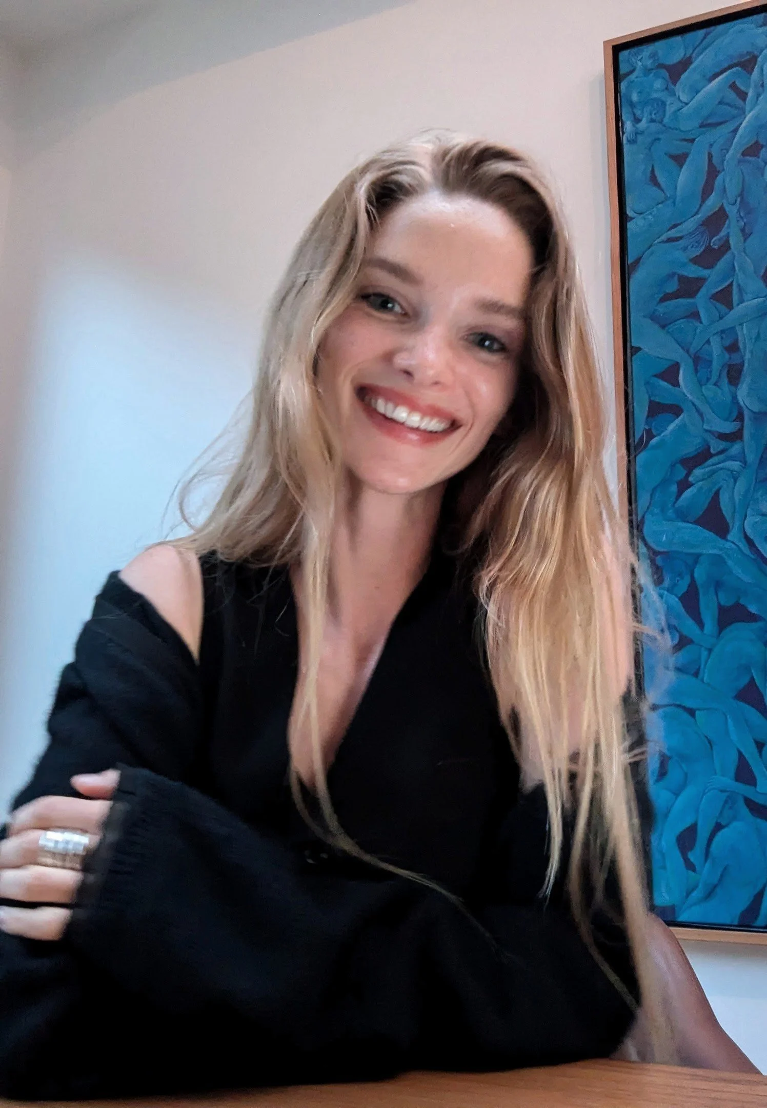 A smiling woman with long blonde hair, wearing a black top with cut-out shoulders, sitting at a table with her arms crossed, against a background of a beige wall with a framed blue abstract artwork.