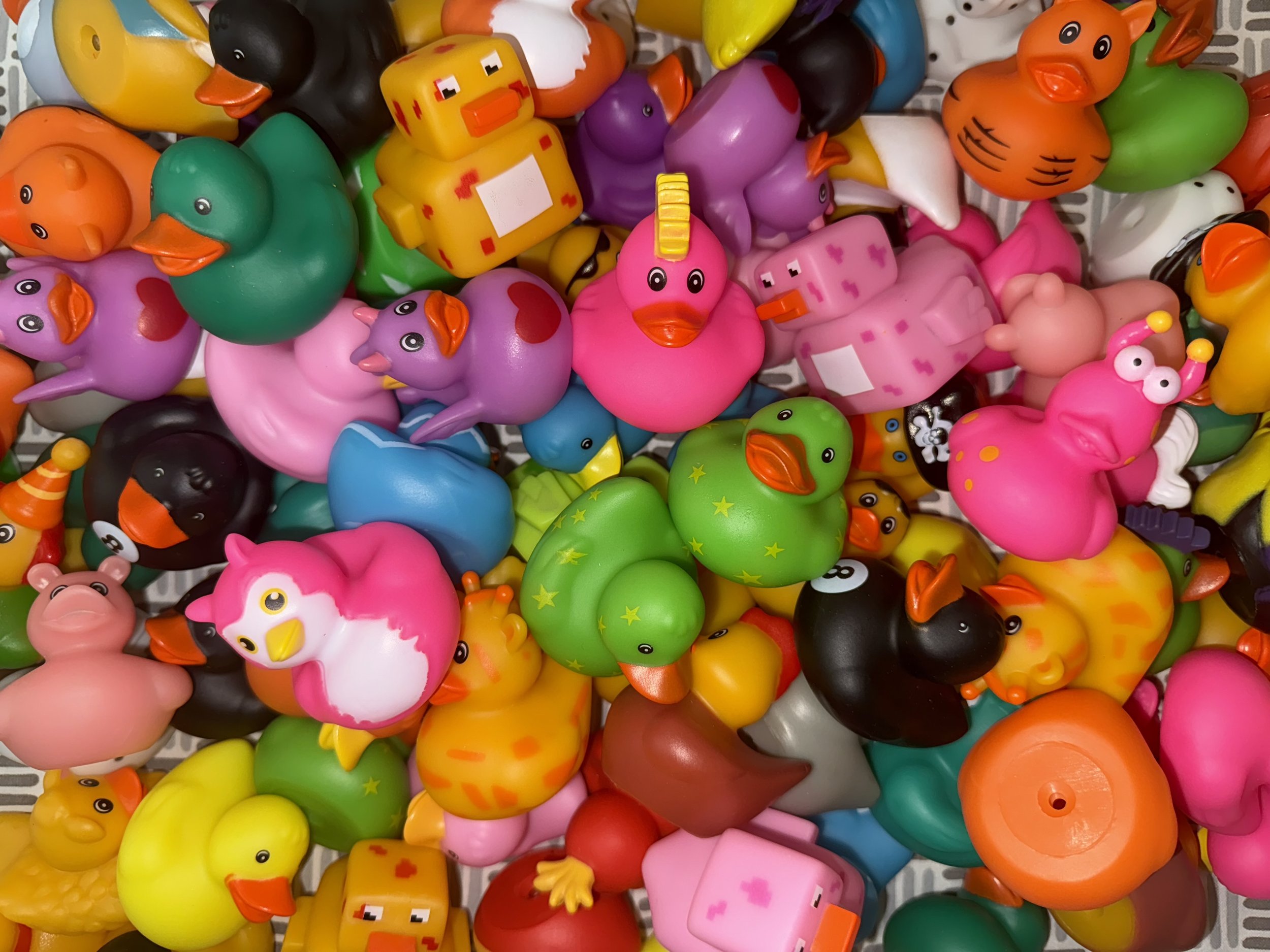 A variety of colorful rubber duck toys pile together, including pink, green, yellow, and orange ducks, with some featuring unique designs like stars and animal costumes.