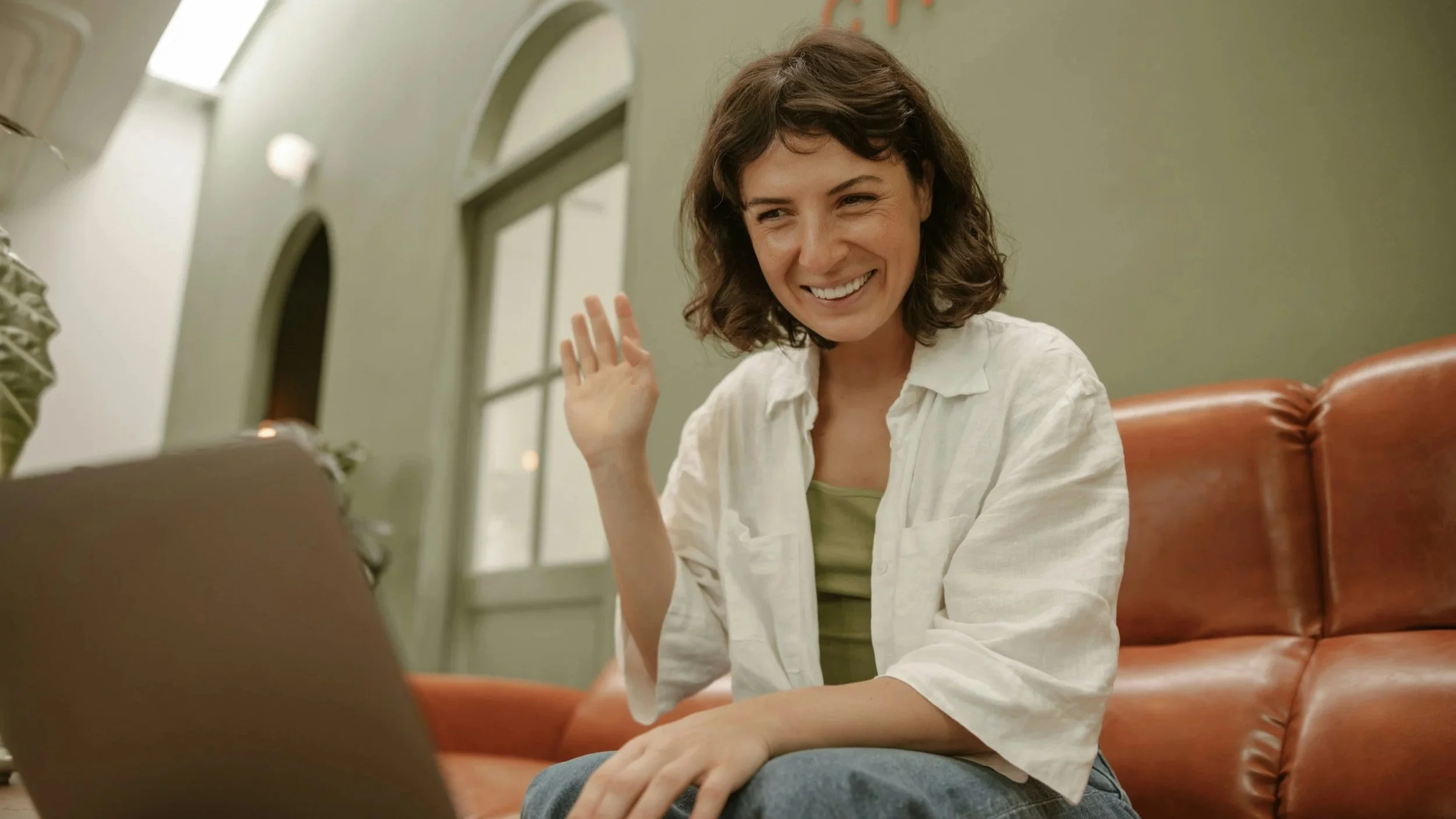 A woman with brown, curly hair smiling and waving at a laptop in a cozy room with large windows and a green wall.