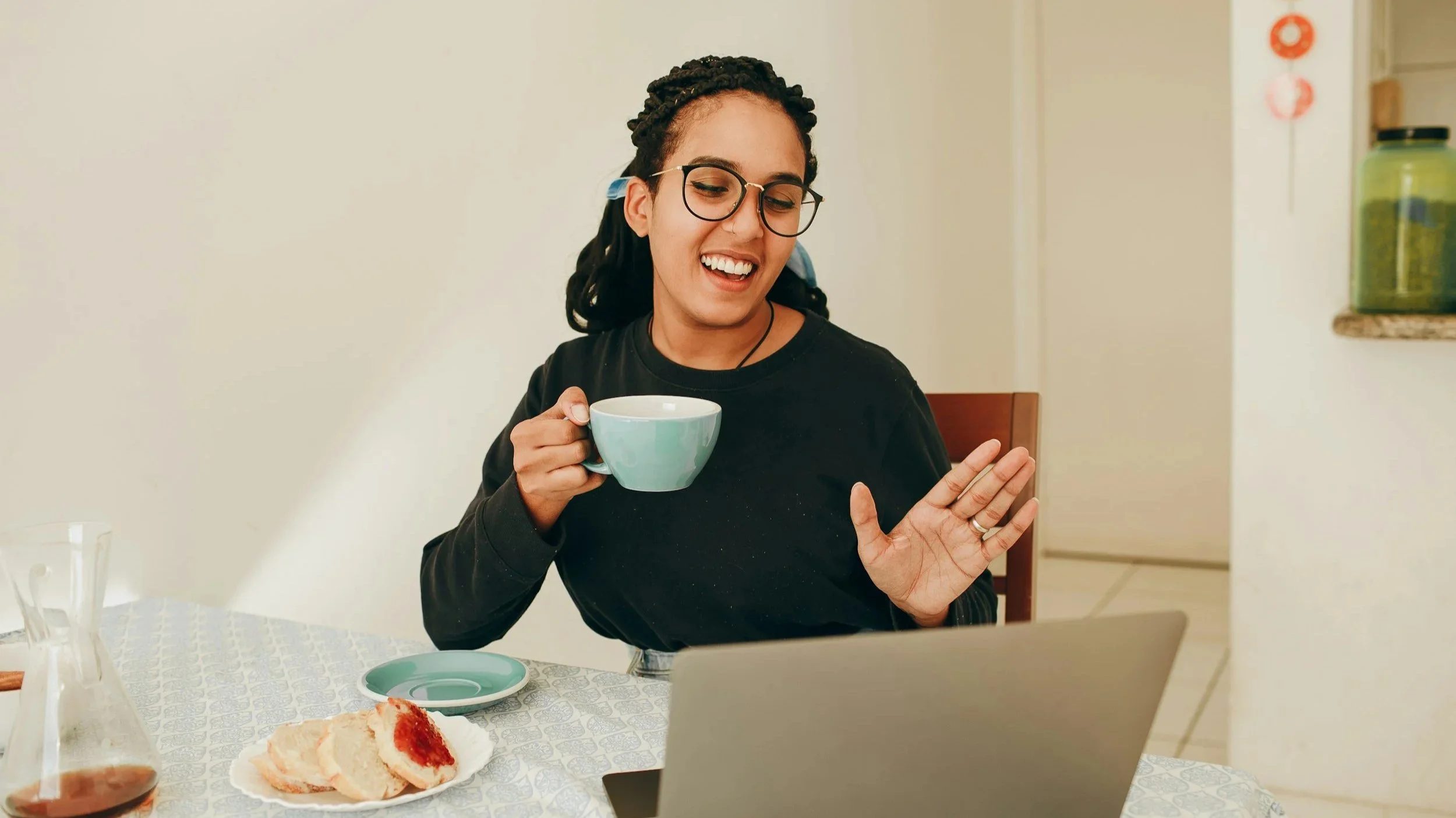 Young woman holding a blue cup, waving at someone on a laptop screen