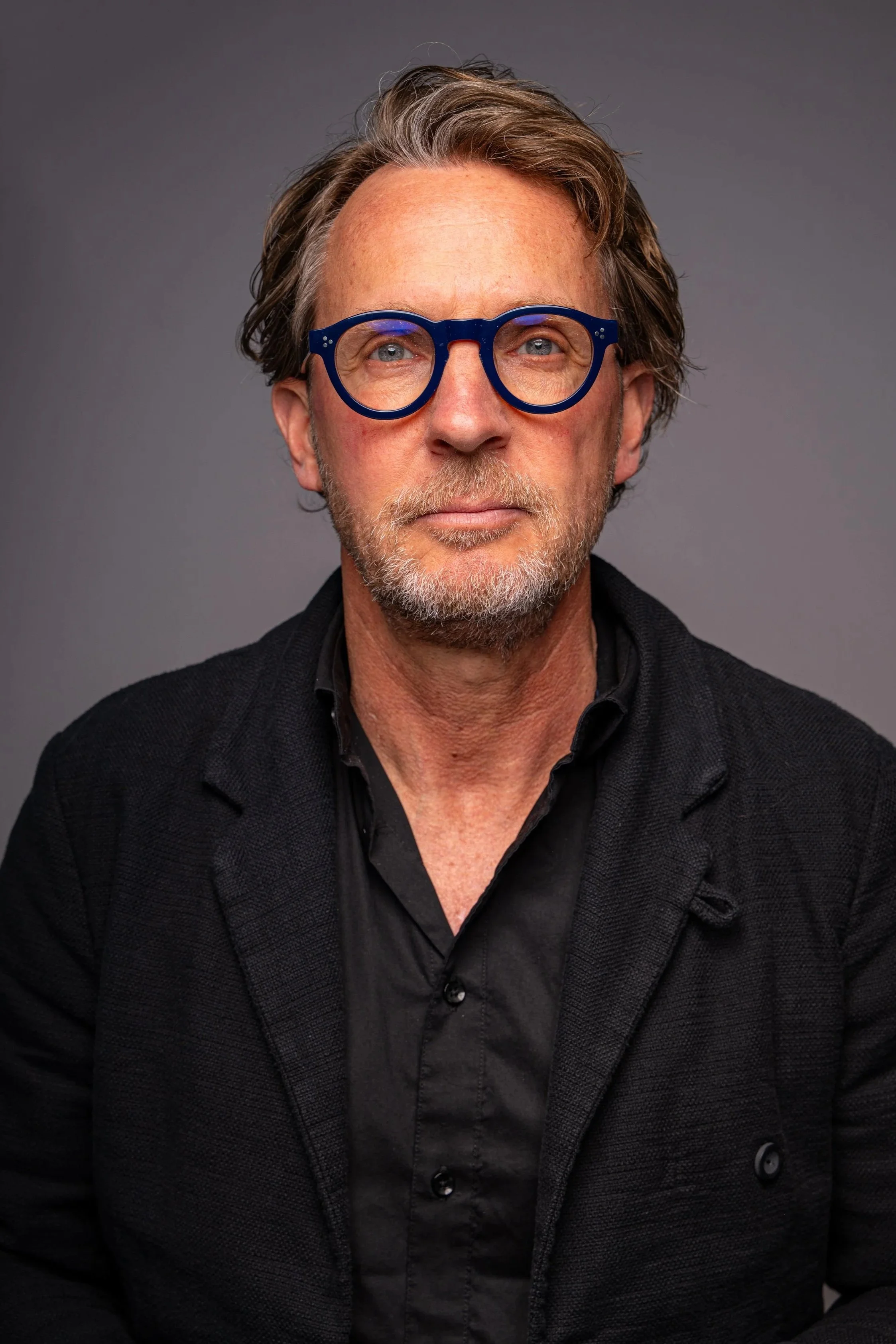 Porträt eines Mannes mit grauem Bart, blauen Augen und braunen, leicht grauen Haaren, trägt blaue Brille, schwarzes Hemd und Jacke, sitzt vor grauem Hintergrund.