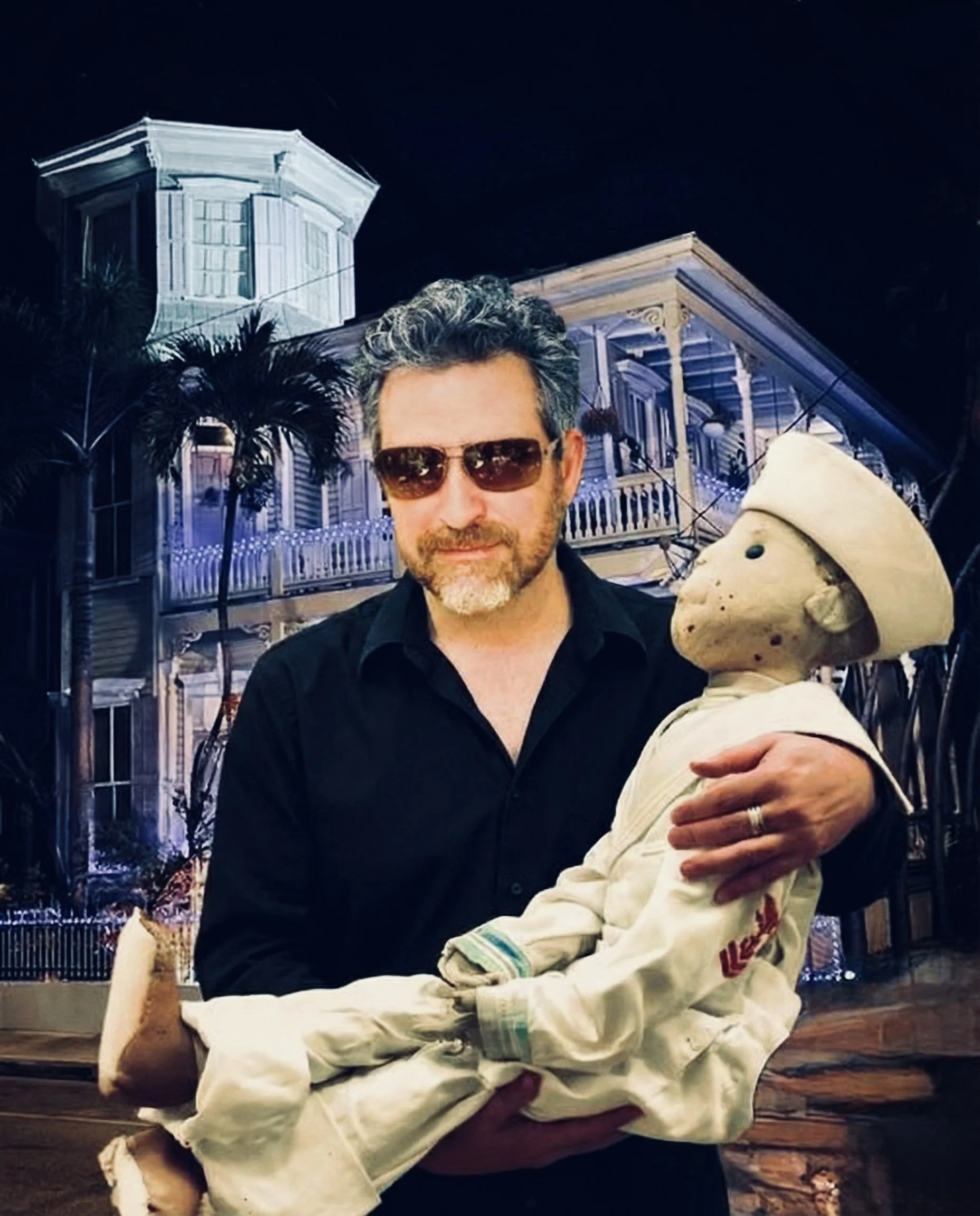 A man with sunglasses, gray hair, and a beard holding a vintage doll in front of a large, lit-up house at night.