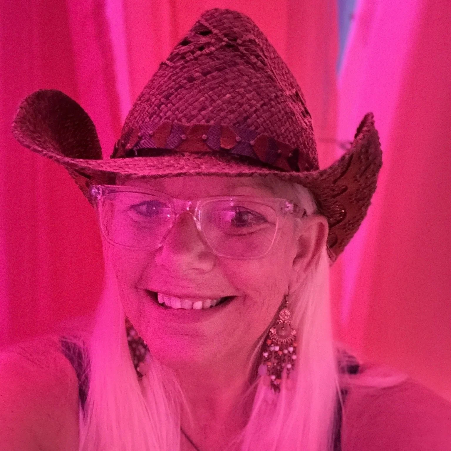 A woman smiling with blonde and a cowboy hat against a pink background.