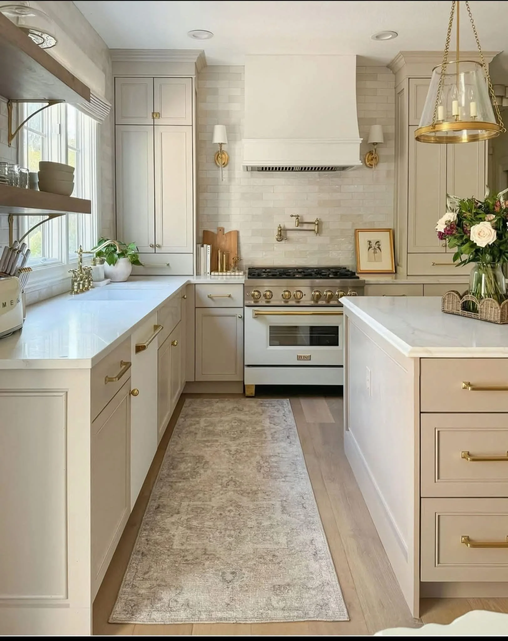 Bright, elegant kitchen with white cabinetry, gold hardware, and a marble island. Features a window, open shelves, a gas stove, and decorative elements like plants and flowers.