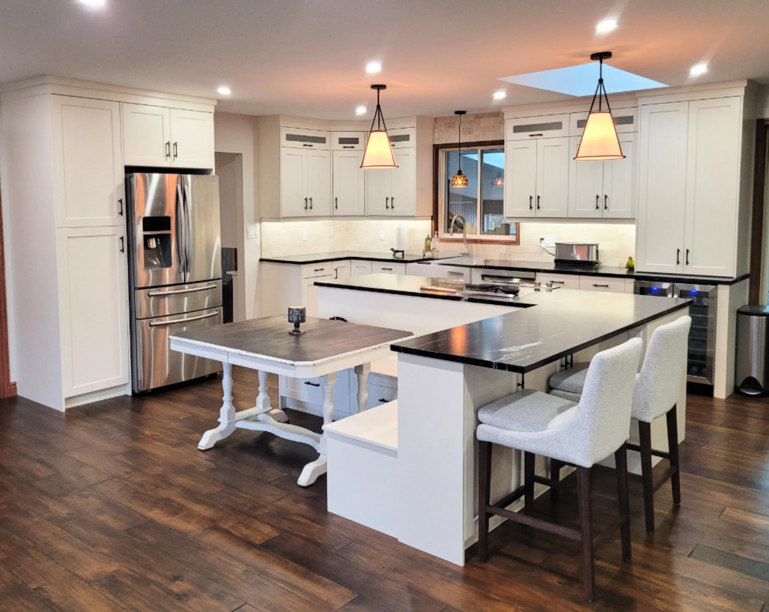 Modern kitchen with white cabinets, stainless steel refrigerator, black countertops, and wooden flooring. Features a kitchen island with seating, a small wooden table, and pendant lights.