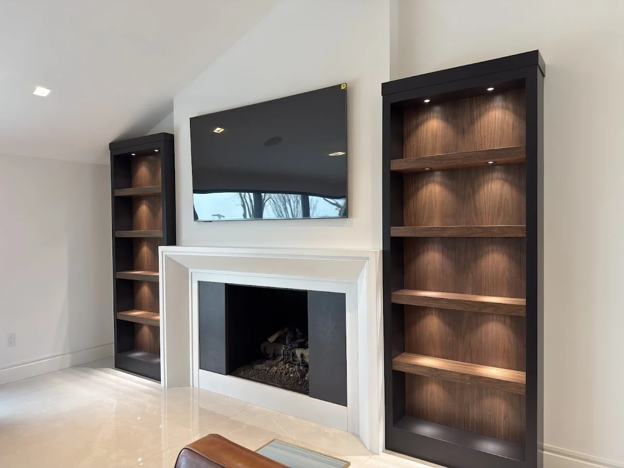 Living room with a wall-mounted flat-screen TV above a fireplace, flanked by two tall black bookshelves with wooden shelves, and a beige tiled floor.