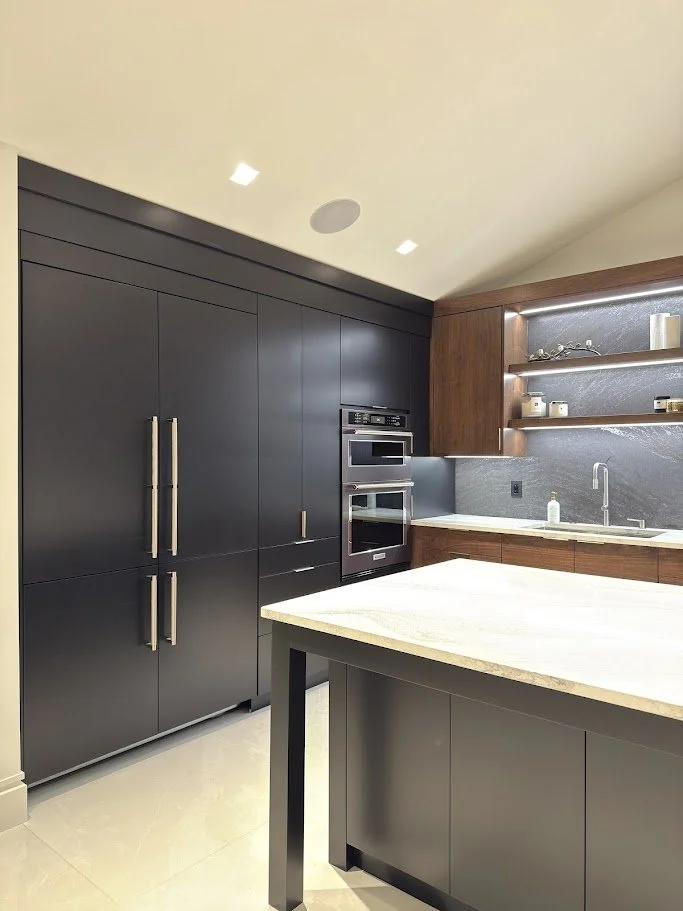 Modern kitchen with black cabinets, built-in oven and microwave, wooden open shelves, and a white marble kitchen island.