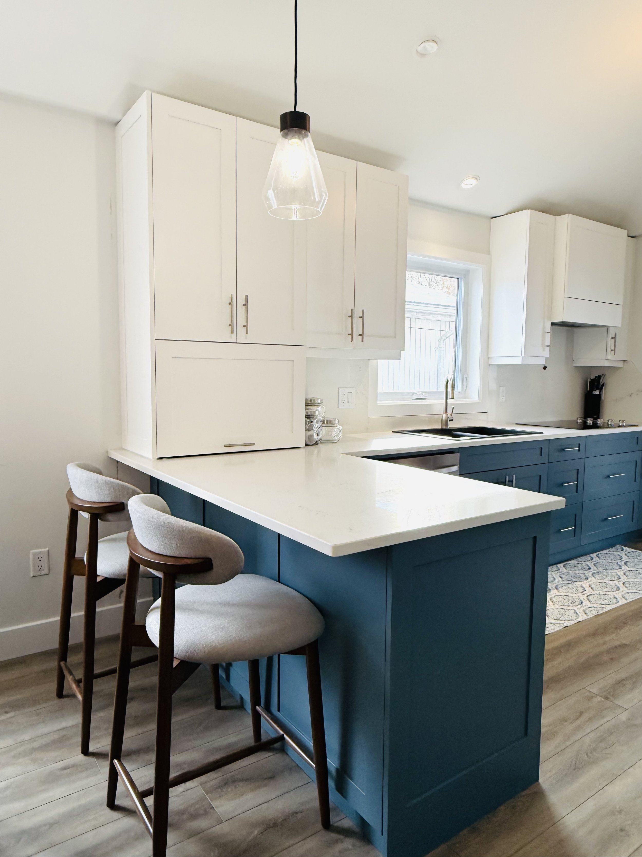 Modern kitchen with white upper cabinets and dark blue lower cabinets, a white countertop, a window over the sink, two beige bar stools, and a hanging light fixture.