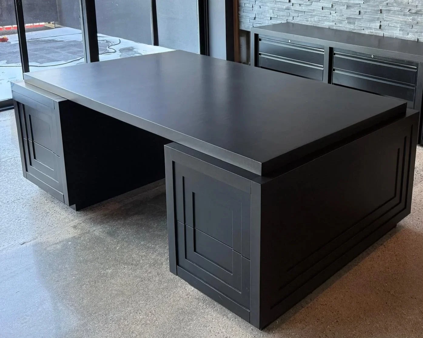 Black office desk with a large rectangular tabletop and storage cabinets on each side, placed in front of a window with a view outside, in a modern office setting.