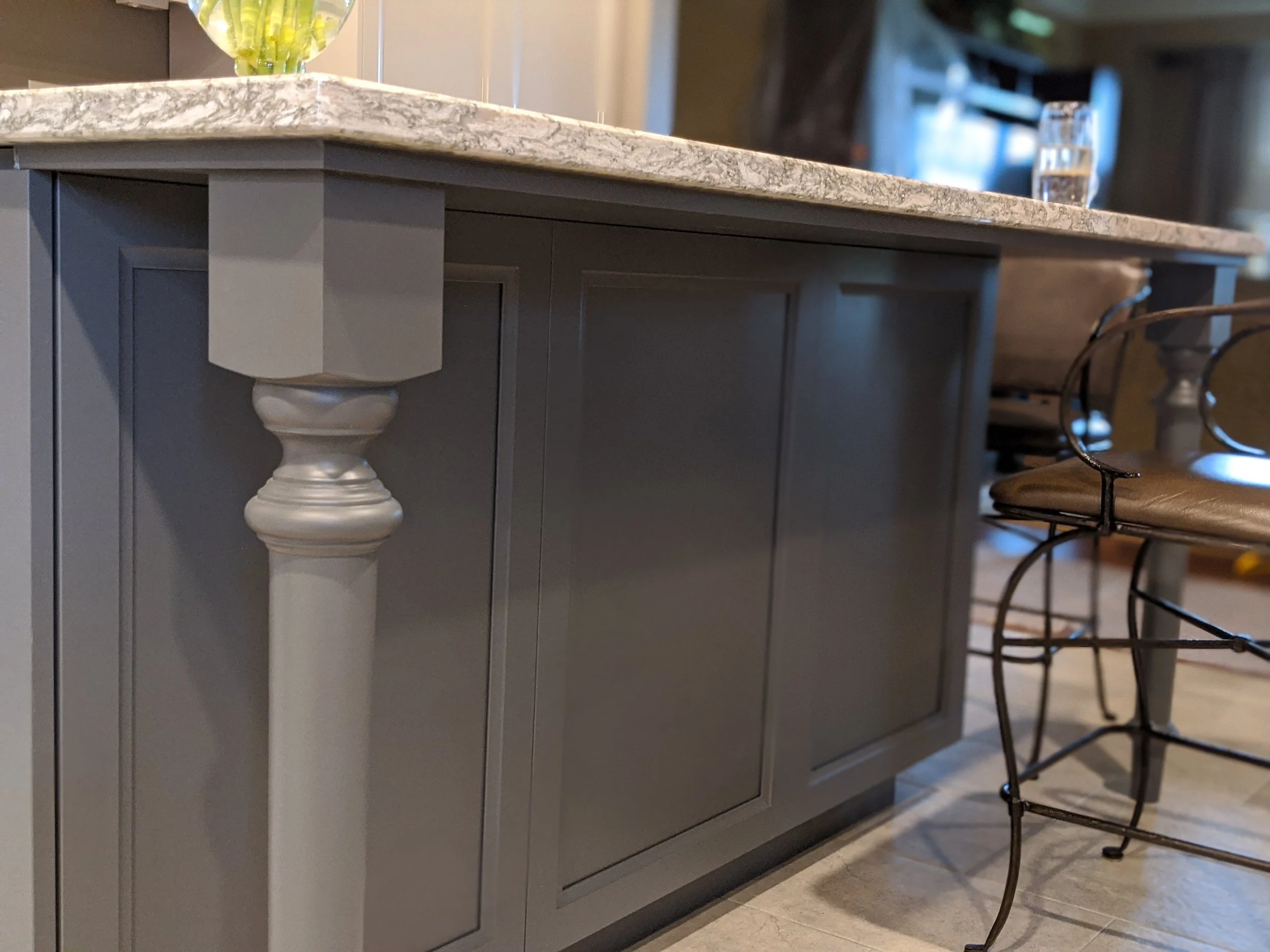 Close-up of a gray kitchen island with a marble countertop and decorative leg, with a glass of water and a yellow fruit bowl on top, and metal chairs with cushioned seats nearby.