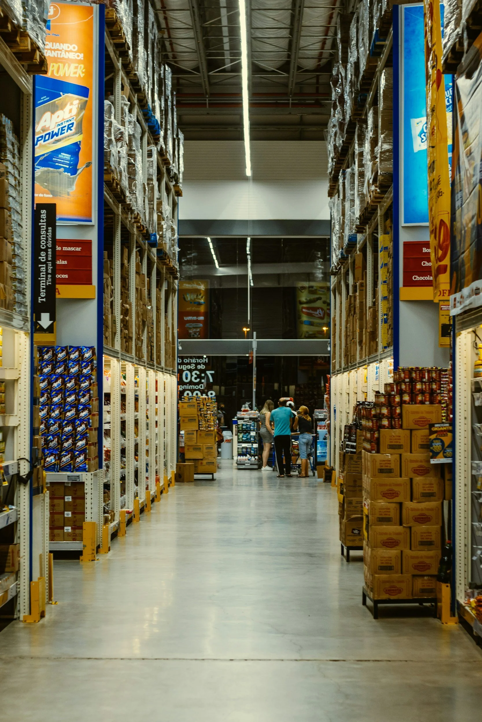 Warehouse aisle with stacked boxes and people shopping.