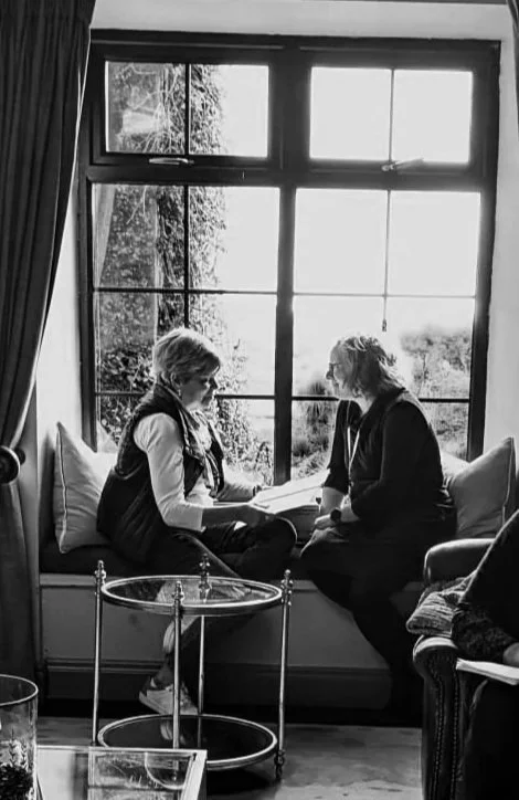 Two women sitting on a window seat and looking at a book, with a large window and curtains behind them.