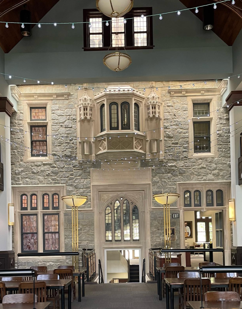 Interior of a public building with a high ceiling, stone wall, and gothic-style windows, featuring hanging string lights, large lamps, wooden tables and chairs, and an ornate fireplace or architectural feature on the wall.