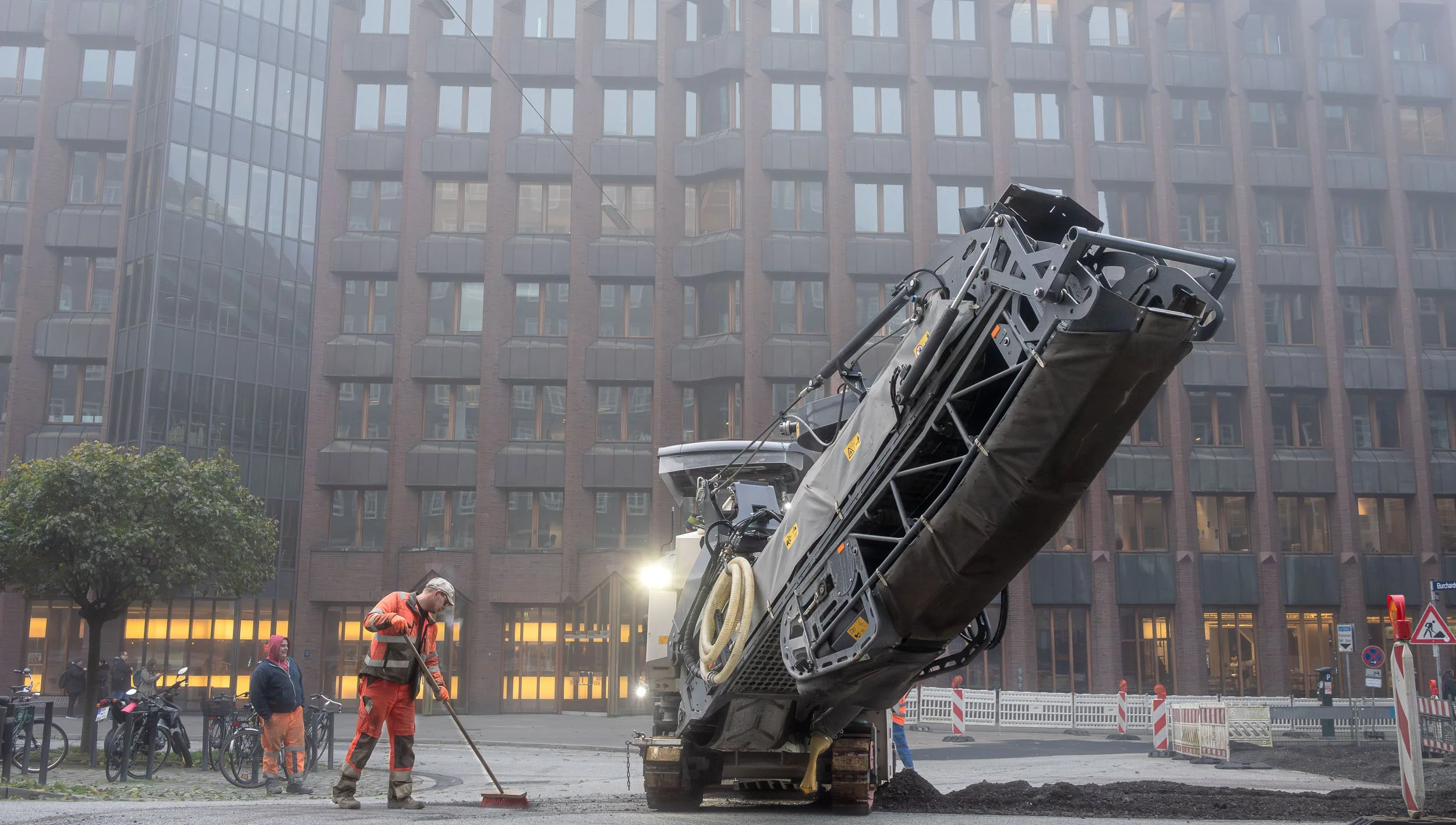 Construction workers operating a ground leveling machine on a city street with a large building in the background.