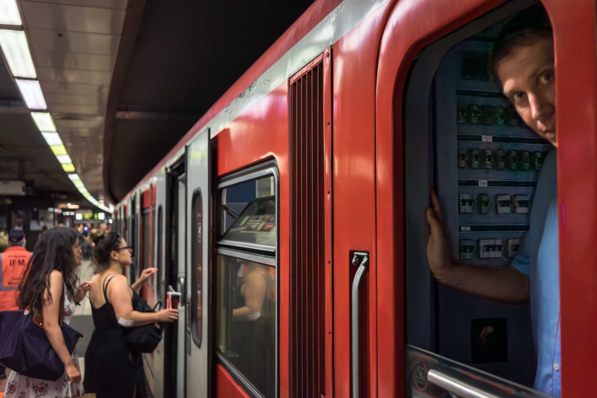 People boarding a red subway train at a station, with one person leaning out of the train window.