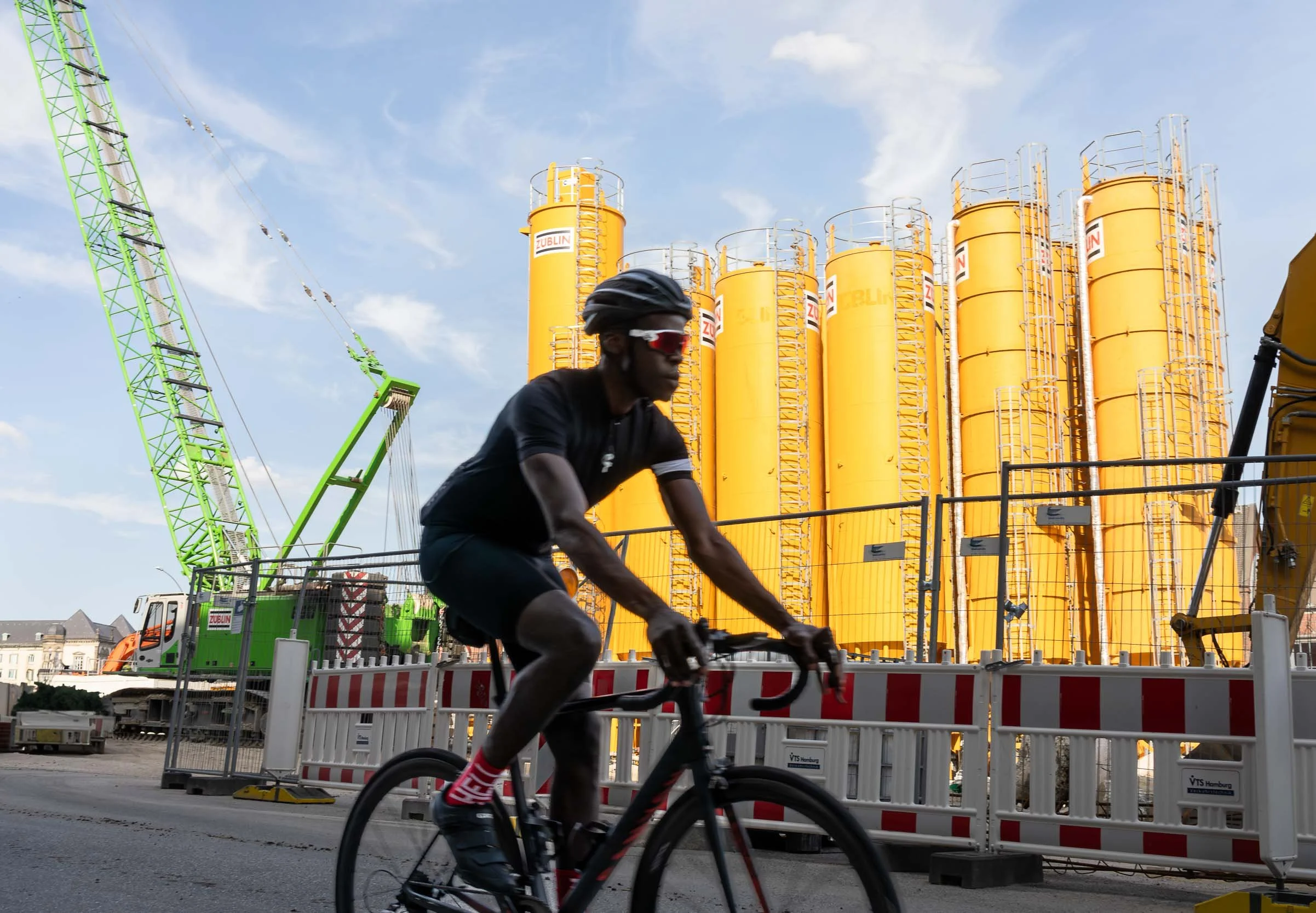 A cyclist riding past a construction site with yellow storage tanks and a green crane.