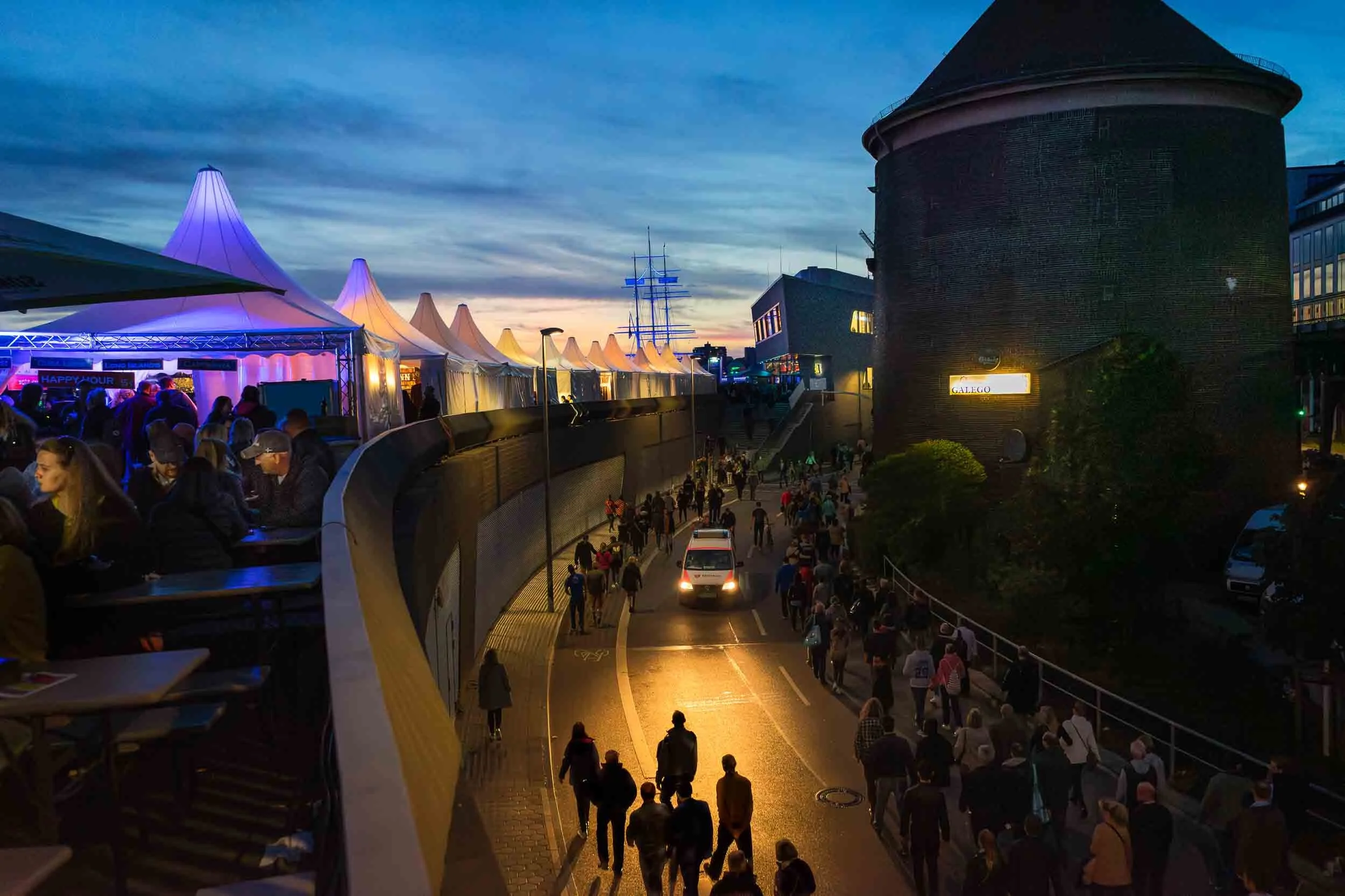 Night scene of a busy street with many people walking, a line of tents with colorful tops on the left, a large brick building on the right, and a ship's mast in the background, under a cloudy sky.