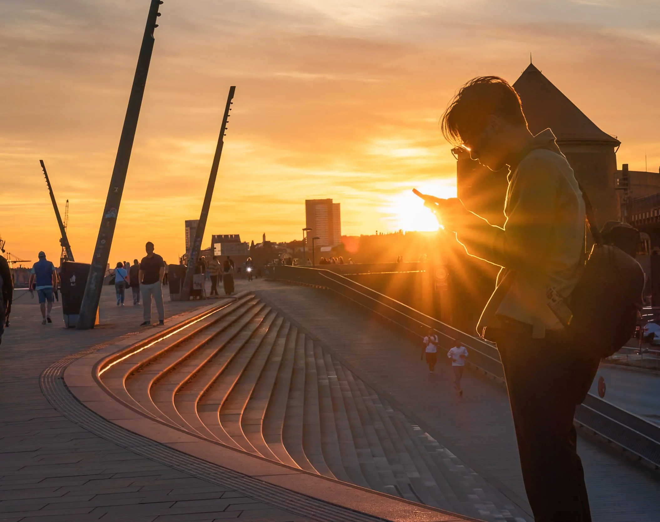 A person using a smartphone during sunset on a city promenade with modern architecture and people walking in the background.