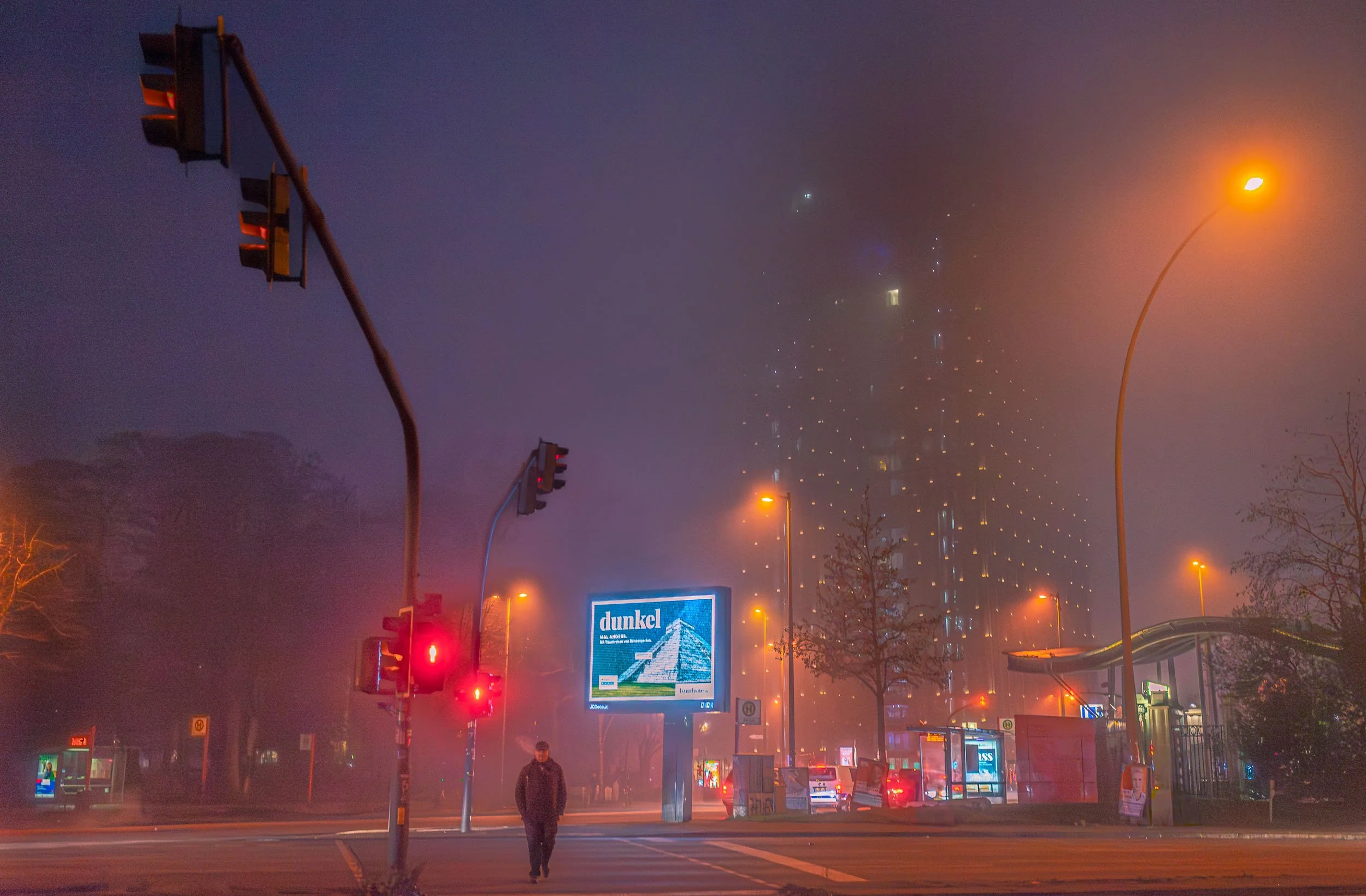 A foggy city street at night with red traffic lights, streetlights, and a tall building in the background. A person is walking at the crosswalk near a bus stop with bright digital advertisements, including one for Dunkel beer.