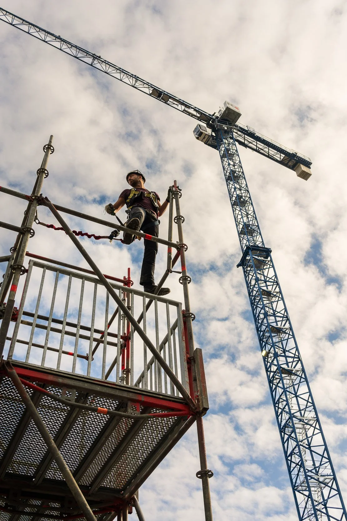 A construction worker standing on scaffolding with a safety harness, next to a tall blue construction crane against a cloudy sky.