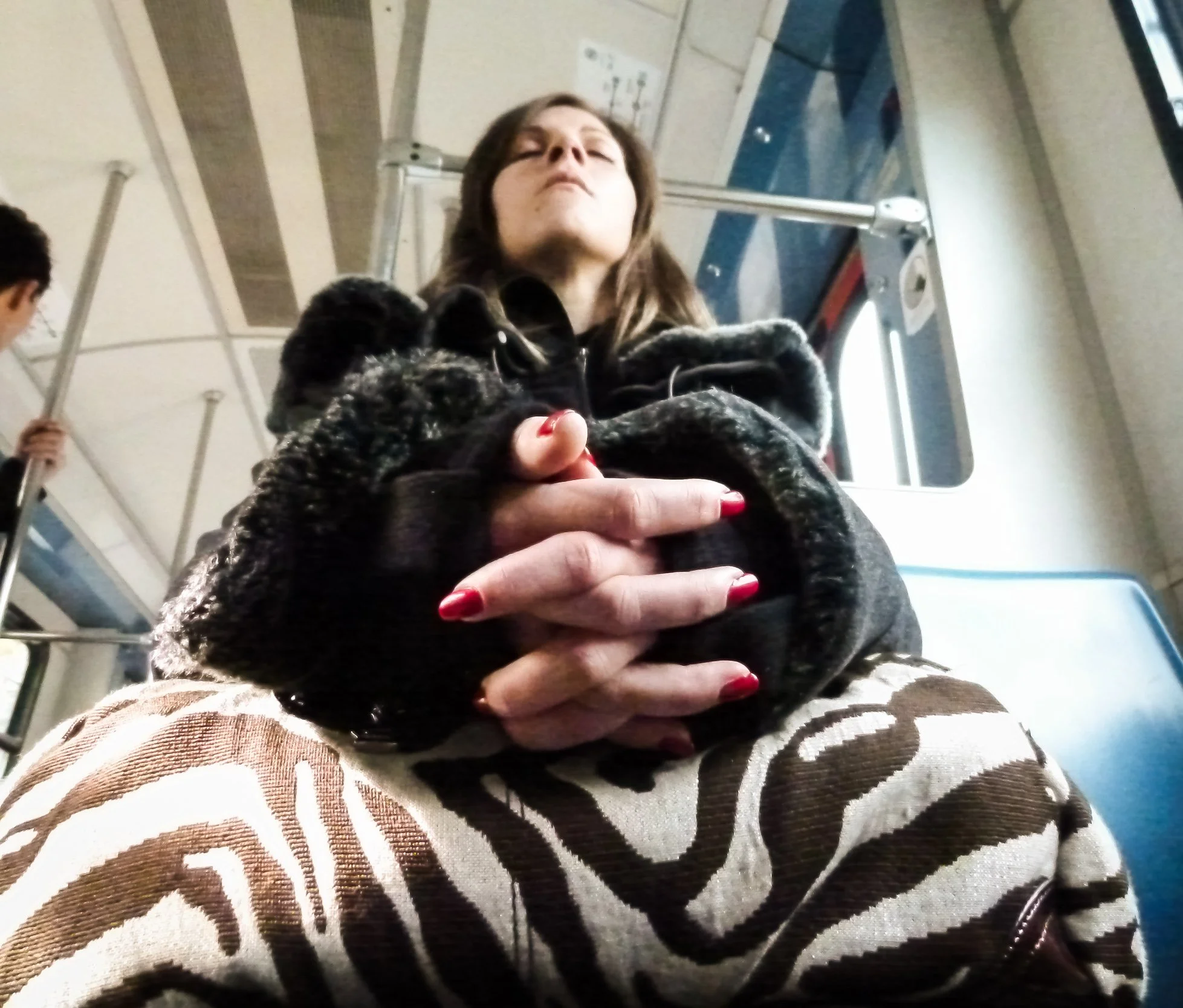 A woman sitting on a bus with her hands clasped, wearing a black fuzzy coat and animal print pants, with a window behind her.