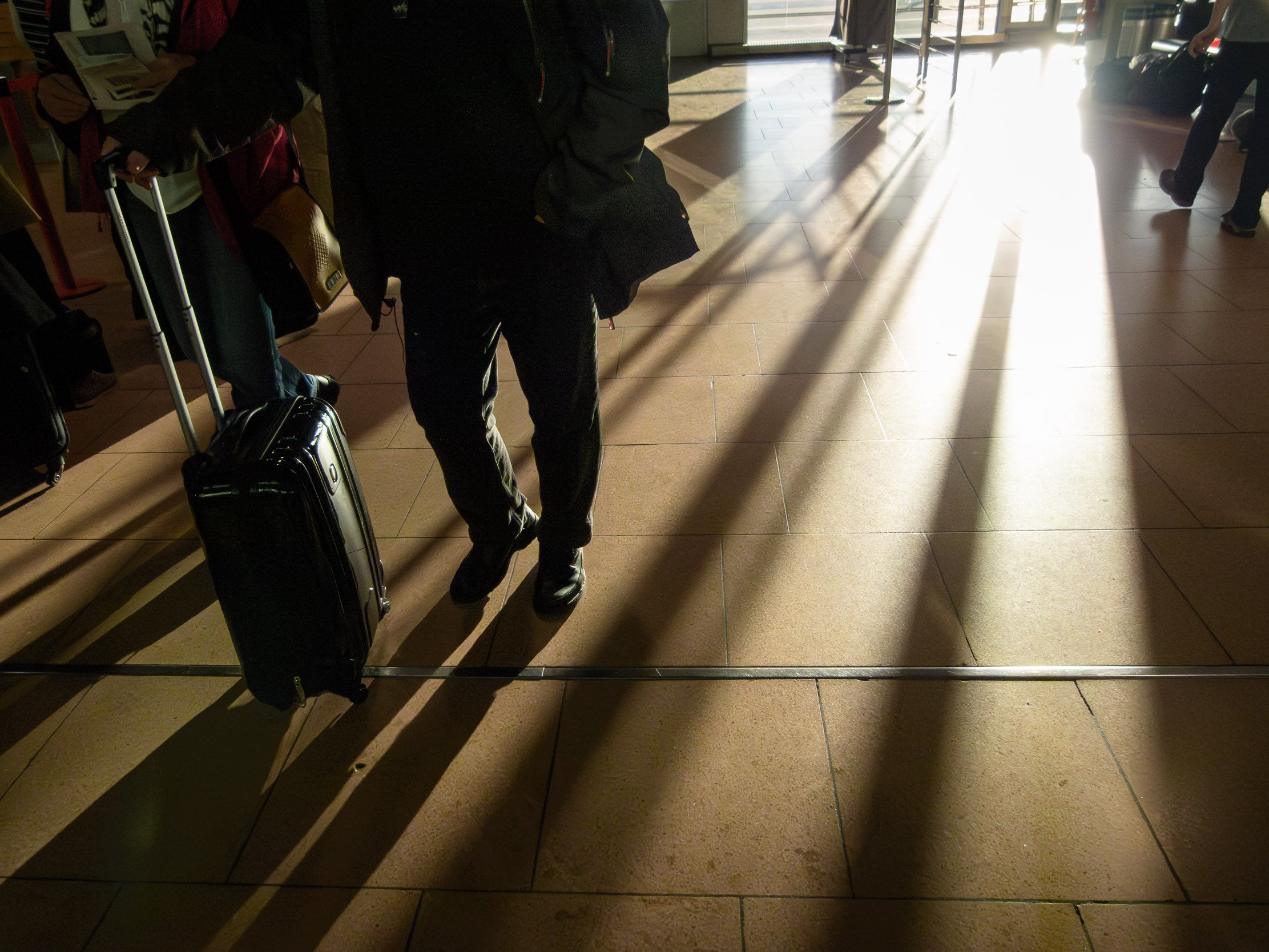 Person standing at airport checking in luggage, with sunlight casting long shadows on the floor.