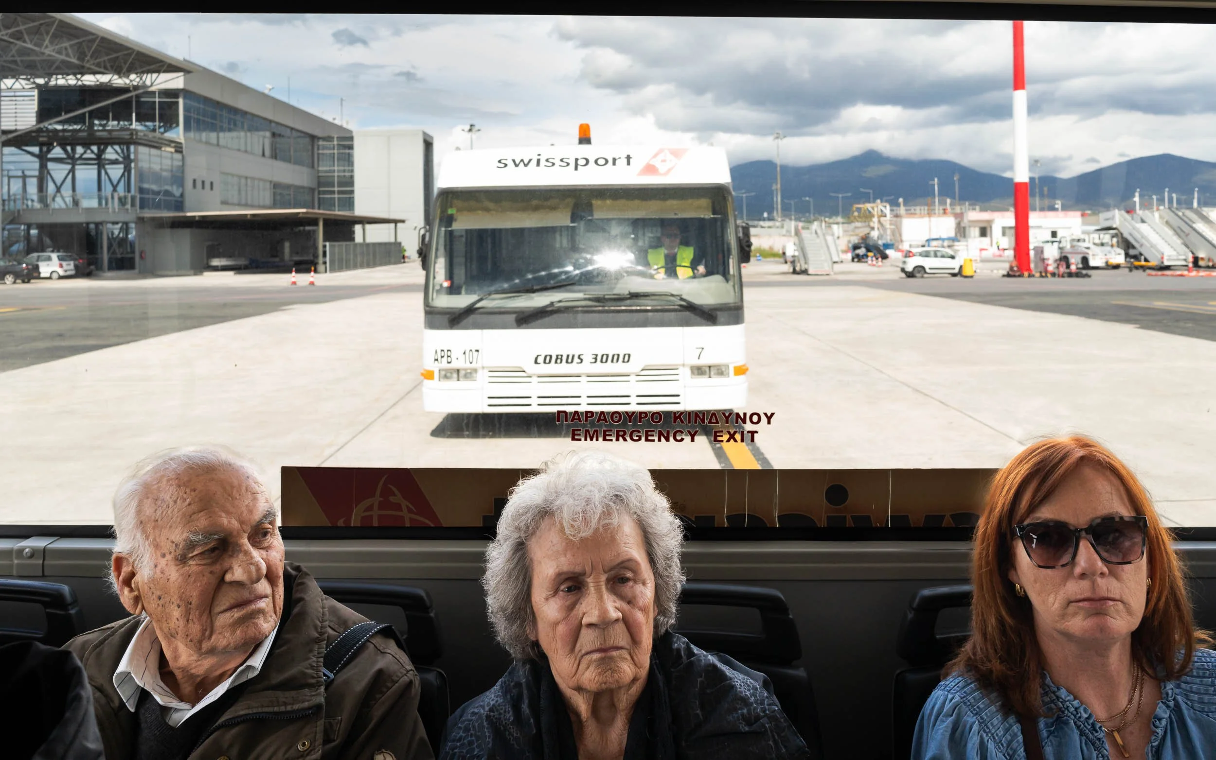 Passengers sitting inside an airport bus, with an aircraft tarmac and a parked bus visible through the window behind them.