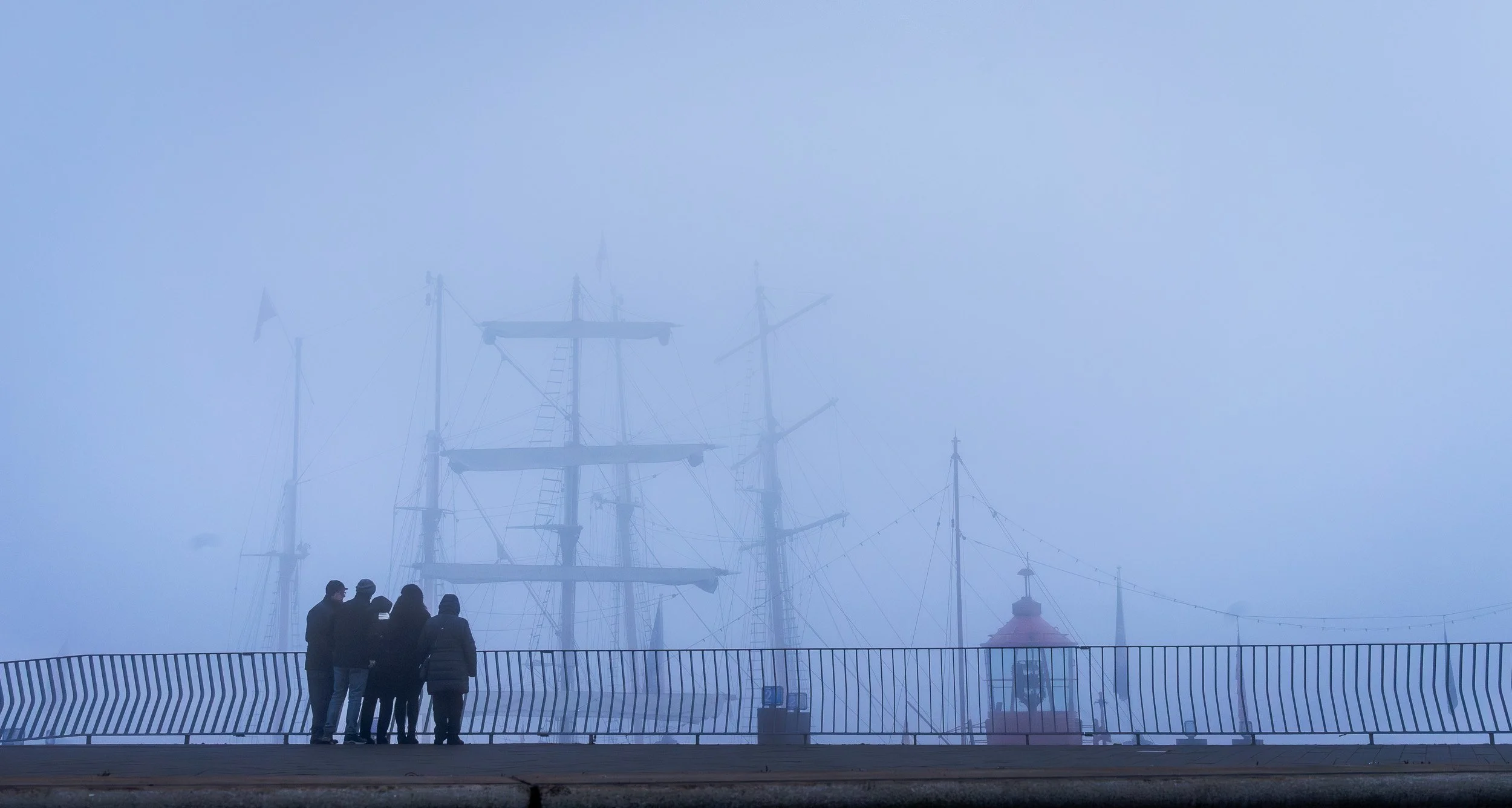 A group of five people standing on a waterfront promenade, looking at fog-shrouded tall ships with masts and rigging, along with a red lighthouse and string lights.