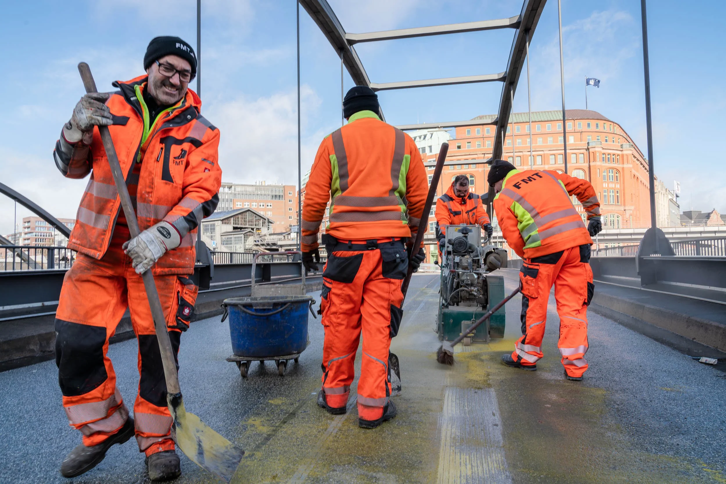 Workers in orange safety gear and helmets are resurfacing a bridge deck with asphalt on an urban bridge, with buildings and a cloudy sky in the background.