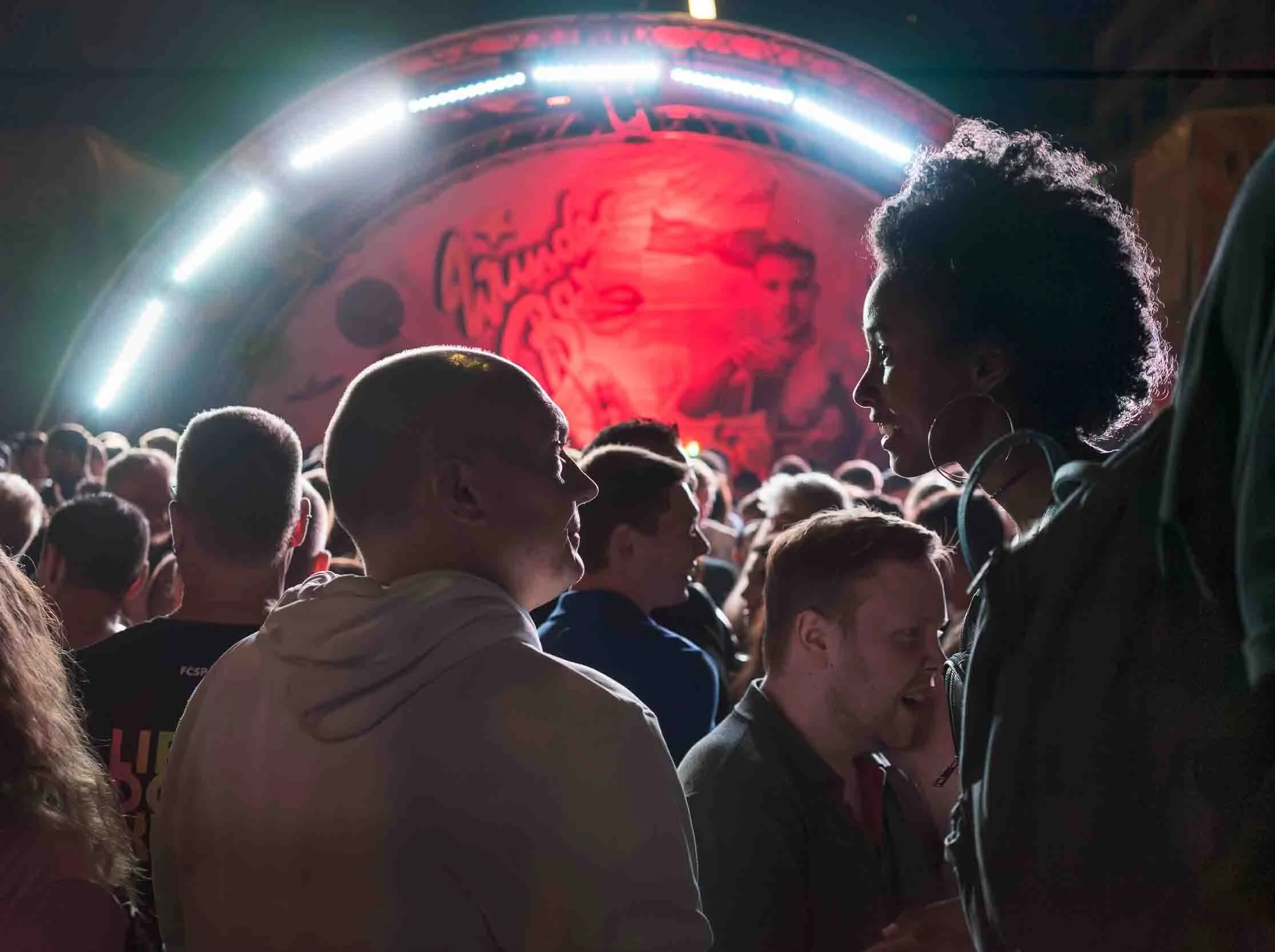Crowd of people at a concert with a large illuminated circular stage in the background displaying a design featuring a face and the word 'LUM' illuminated in red.