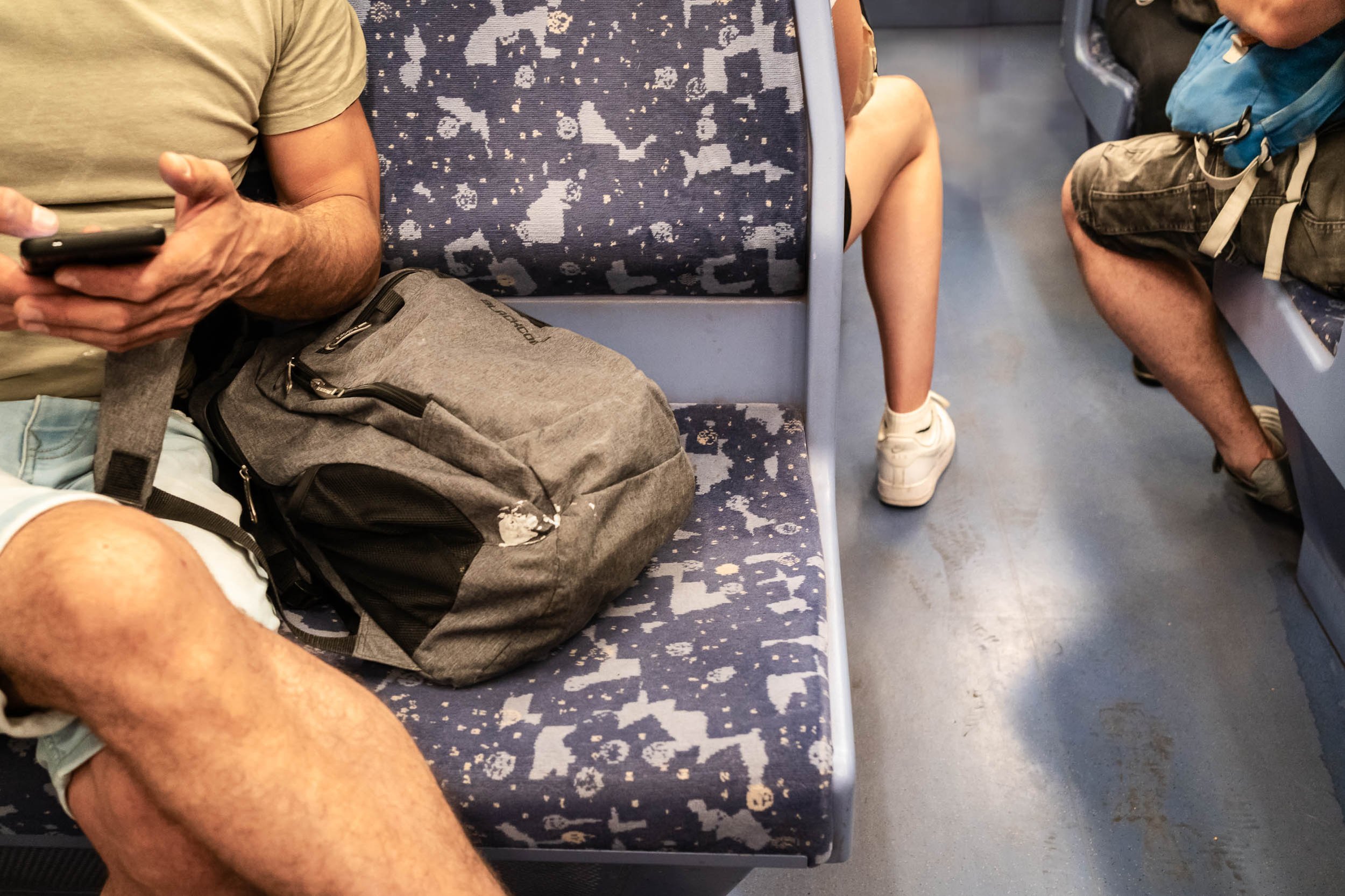 People sitting on a bus, showing a backpack, a person with a phone, and a person with a backpack. The seats have a blue patterned upholstery, and the floor is a grayish color.