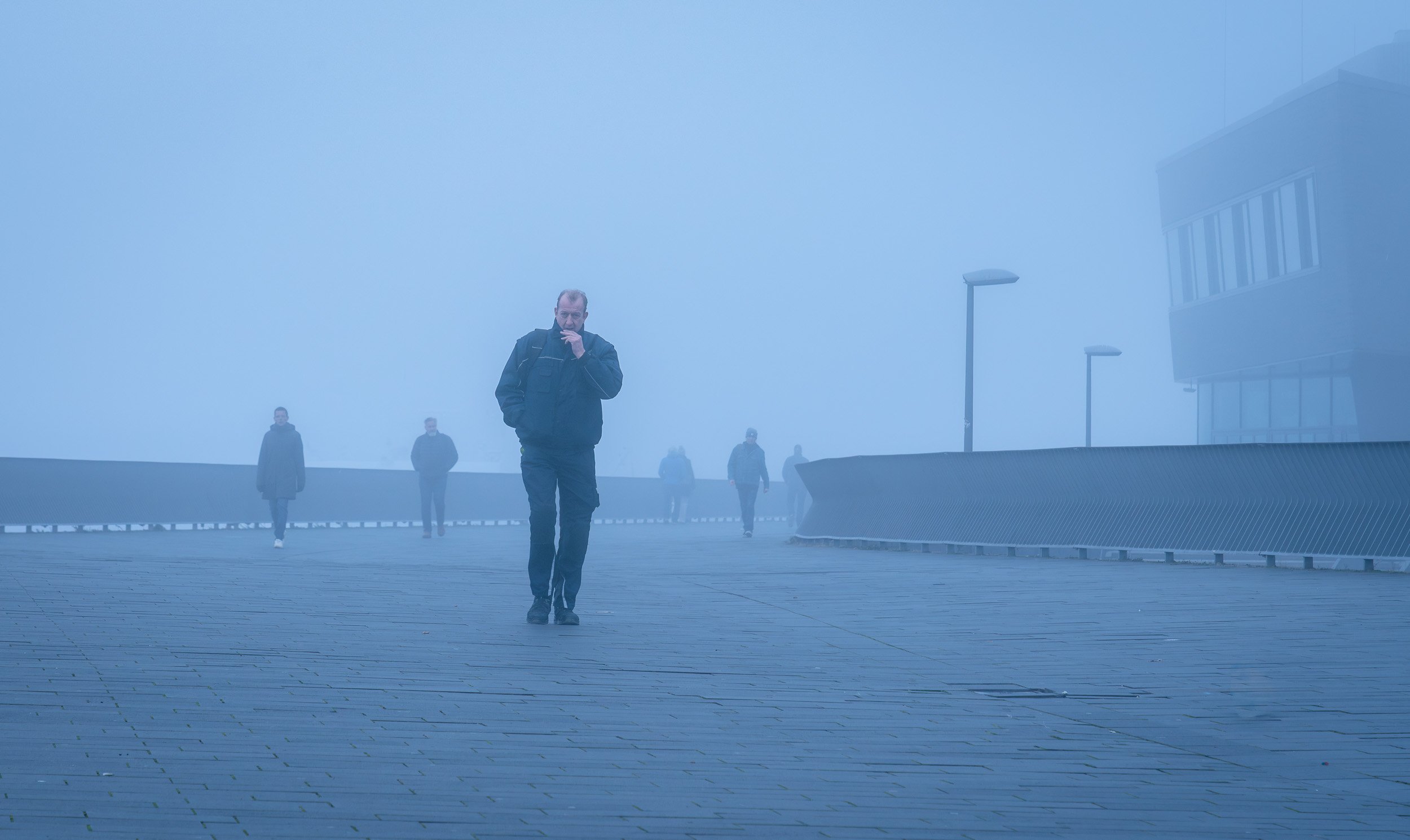 Group of people walking on foggy outdoor walkway, with one man in the foreground and others in the distance, misty cityscape background.