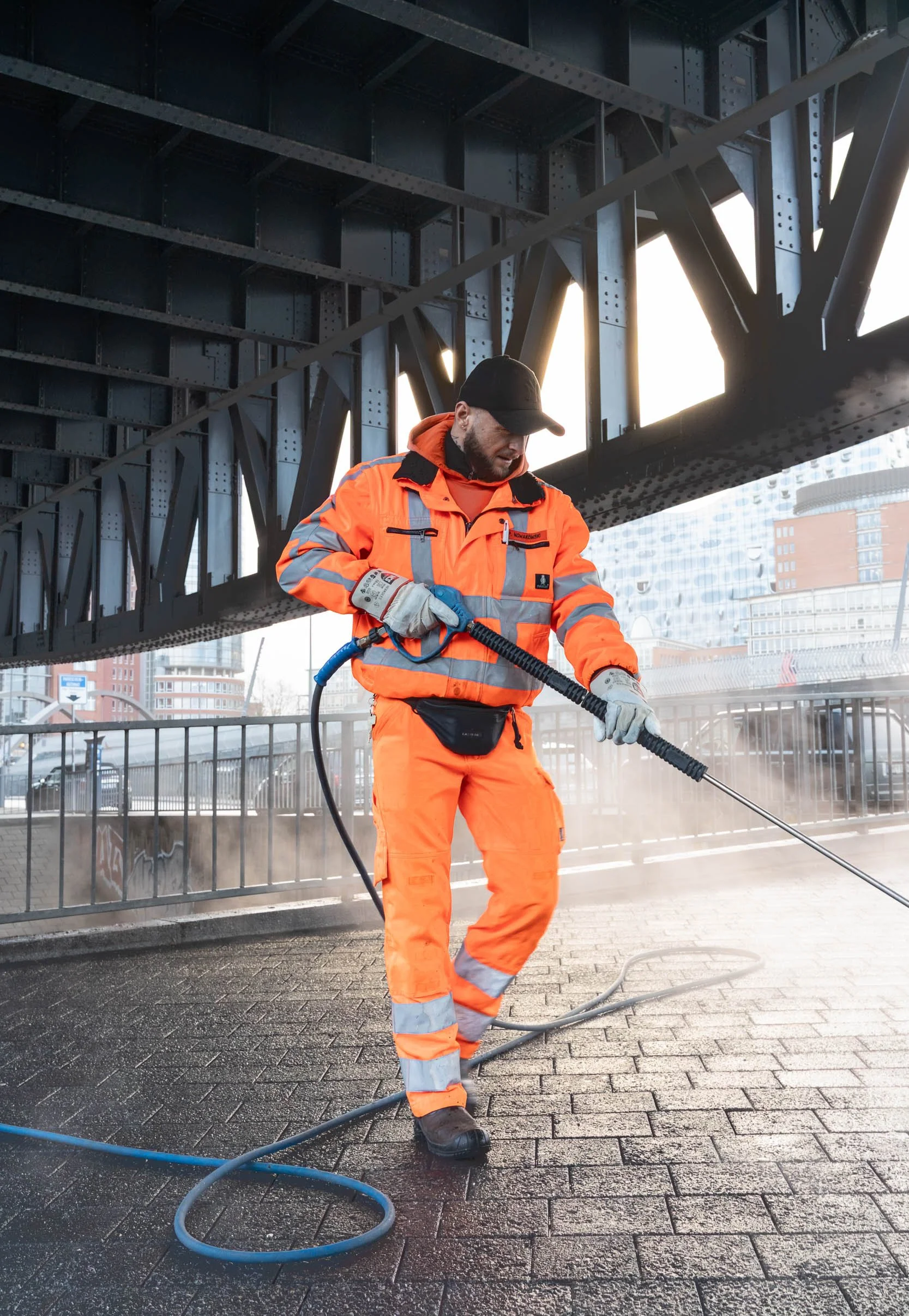 A worker in orange safety gear pressure washing pavement under a bridge in an urban area.
