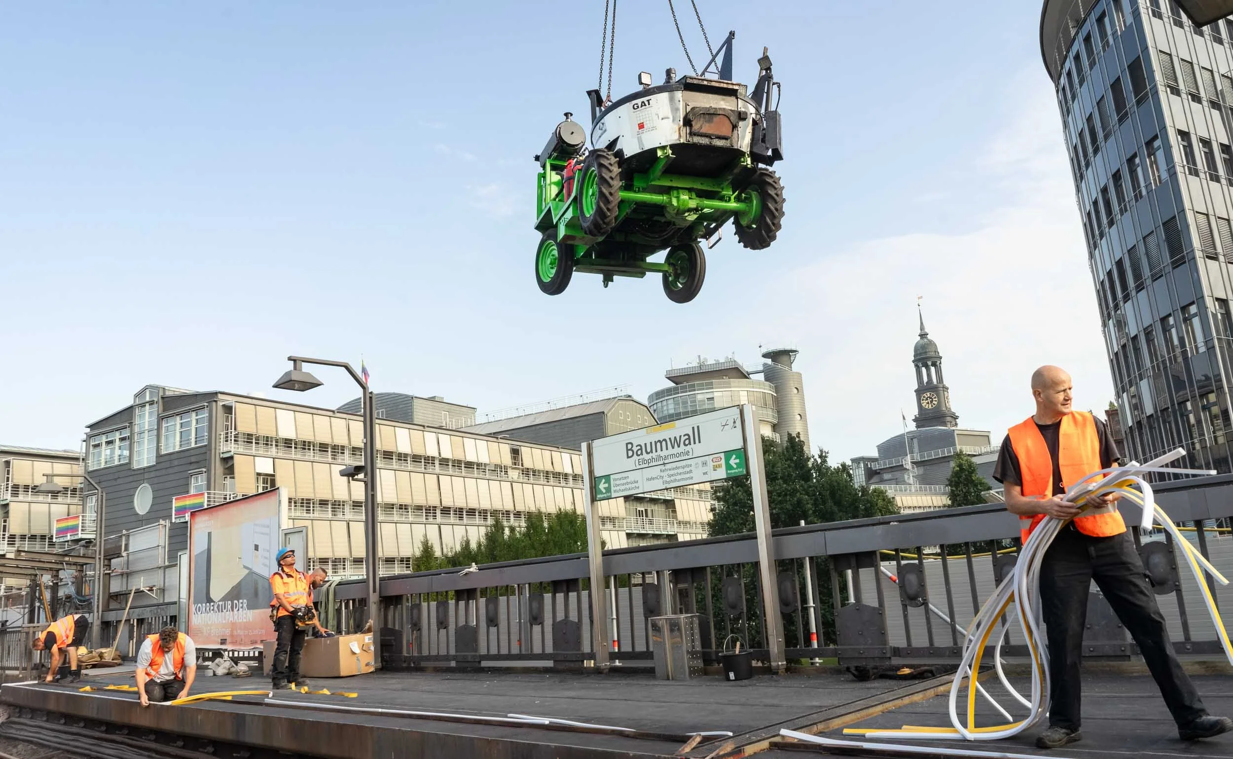 People working on a construction site with a cityscape background. A small vehicle is suspended in the air above them, and they are handling cables and tools.