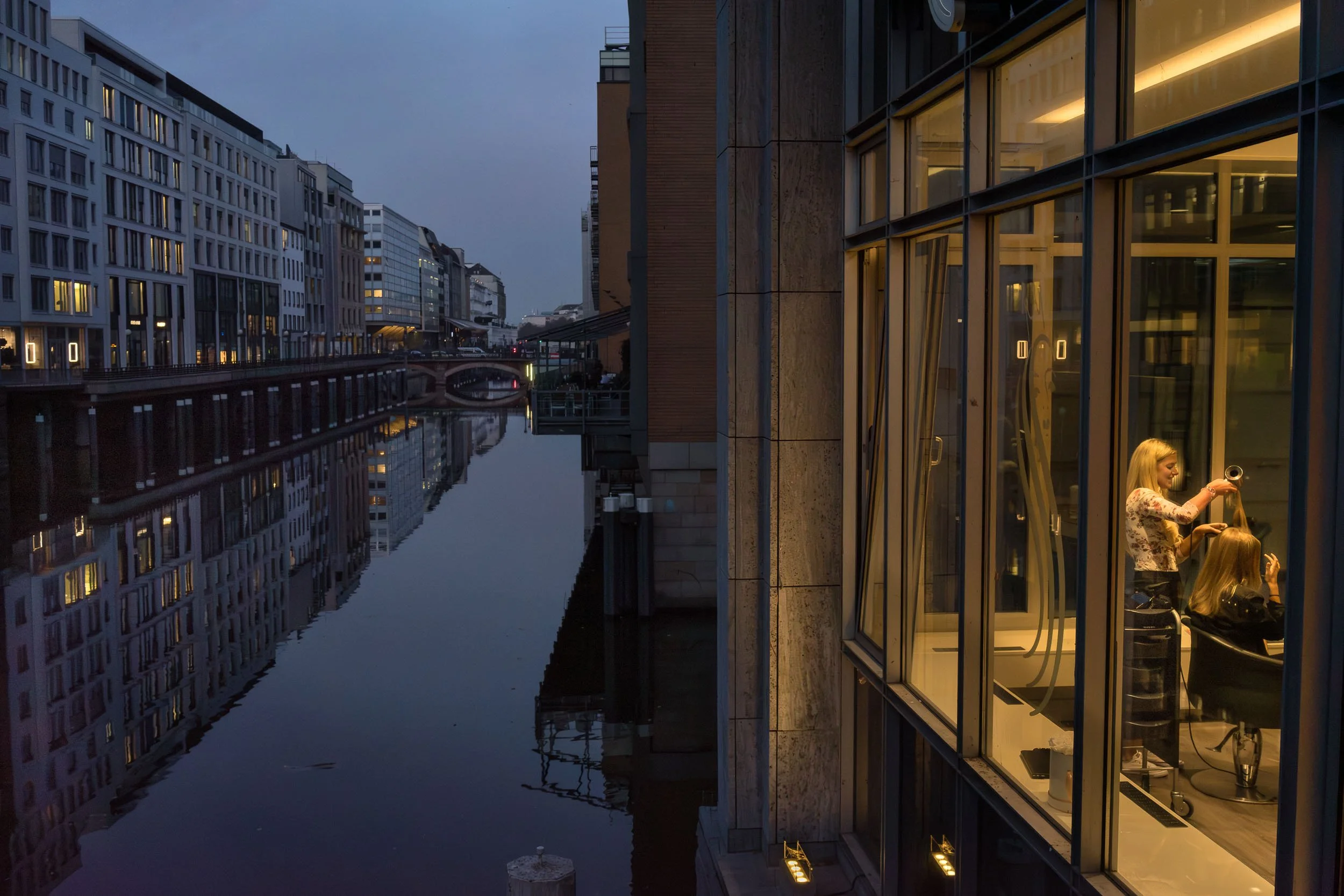 A woman getting her hair styled in a hair salon with large windows overlooking a canal in an urban area at dusk.