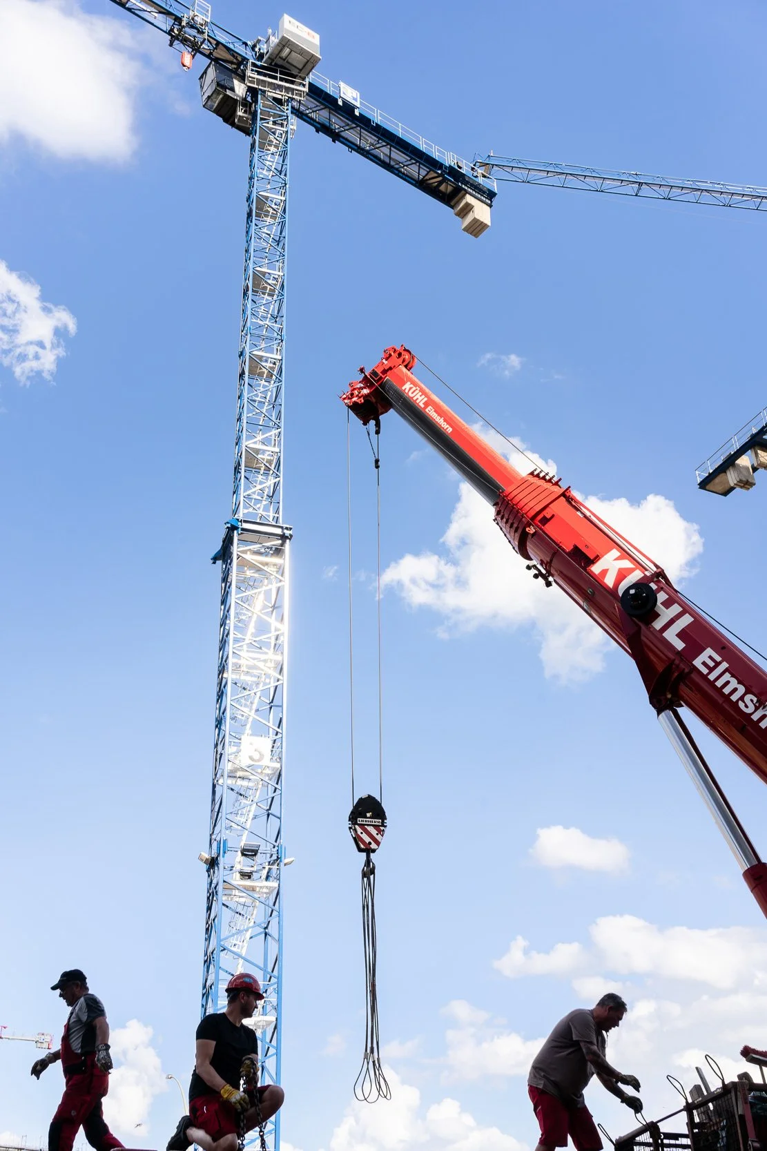 Construction workers are working with a large blue tower crane and a red mobile crane on a construction site under a blue sky with clouds.