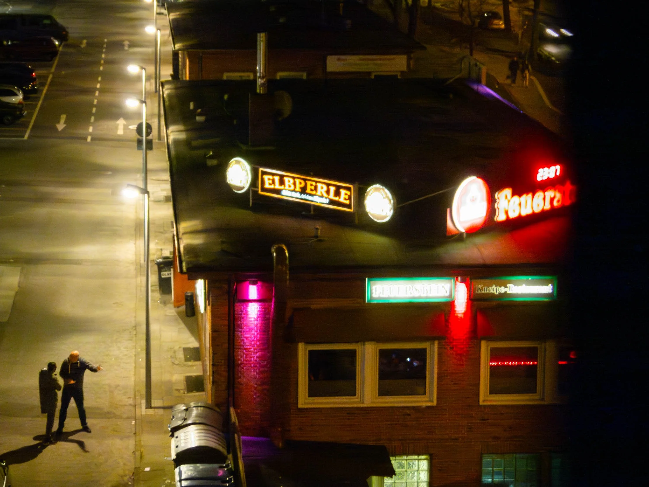 Nighttime view of a restaurant with illuminated signs. Two people are standing on the sidewalk near parked cars, and streetlights light the street.