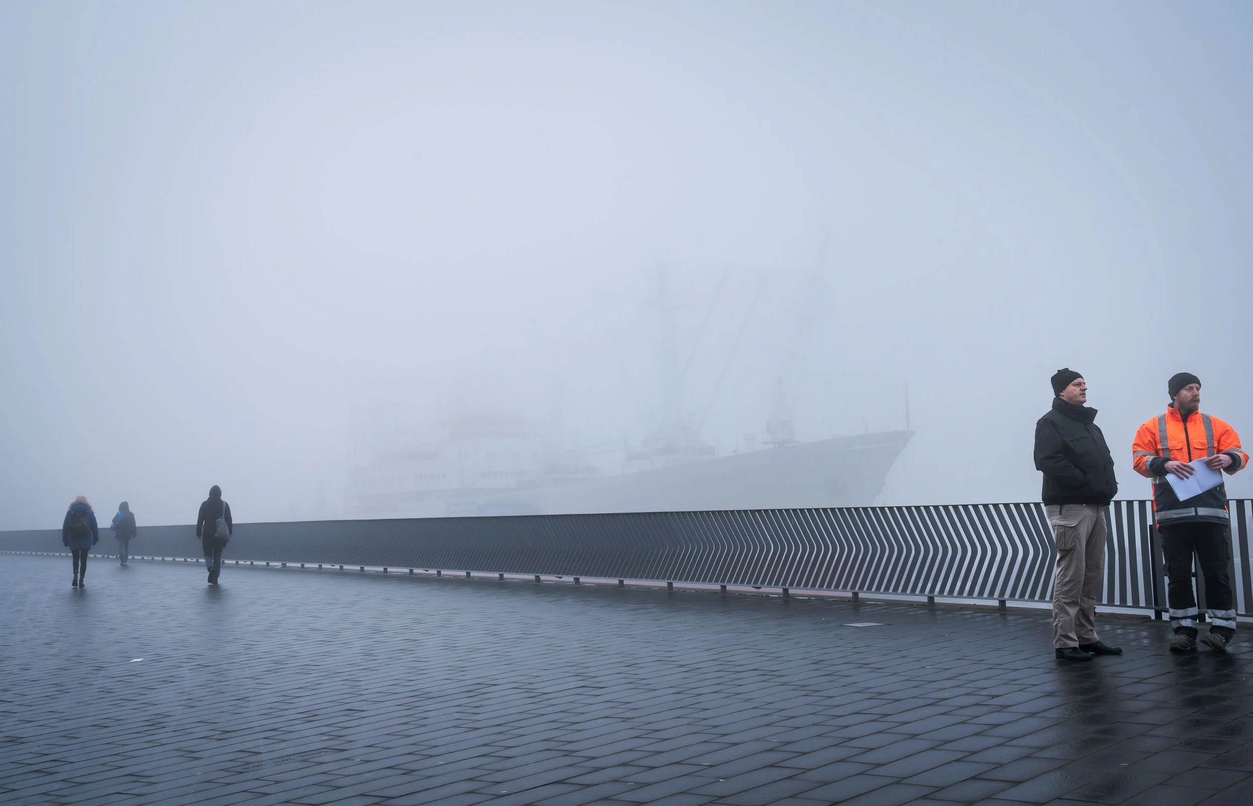 Four people walking or standing on a foggy waterfront promenade with a modern railing, with a faintly visible large ship in the background.