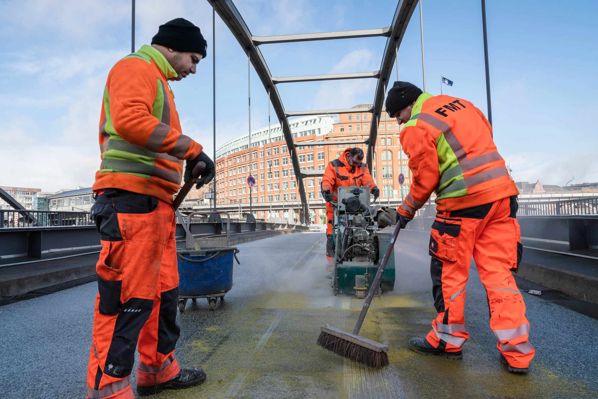 Three construction workers in orange and yellow safety gear working on a bridge surface with a paving machine in the background, city buildings visible behind.