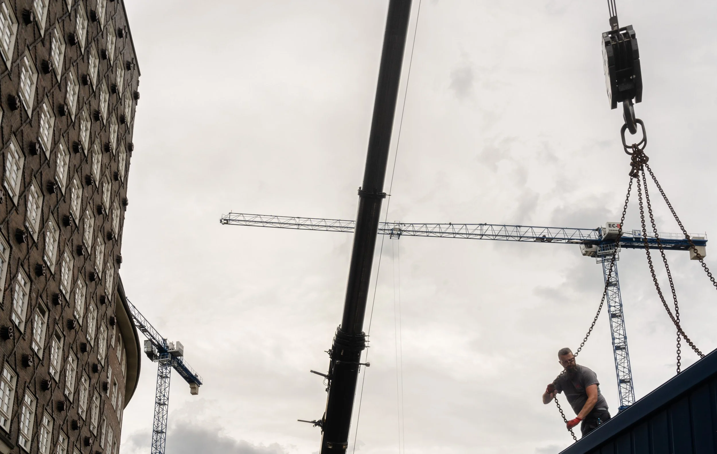 A construction worker operating a crane in an urban area, with tall buildings and multiple crane structures visible against a cloudy sky.