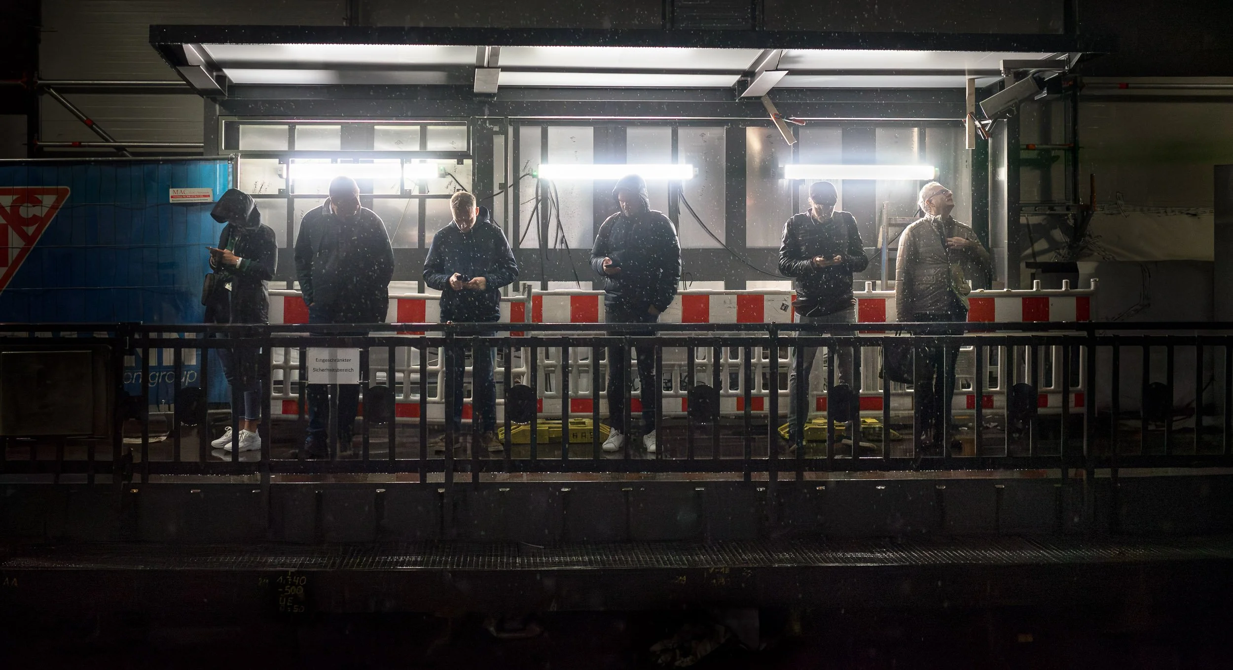 Six people standing on a platform next to a train, looking at their phones, with bright lights above them and rain falling.
