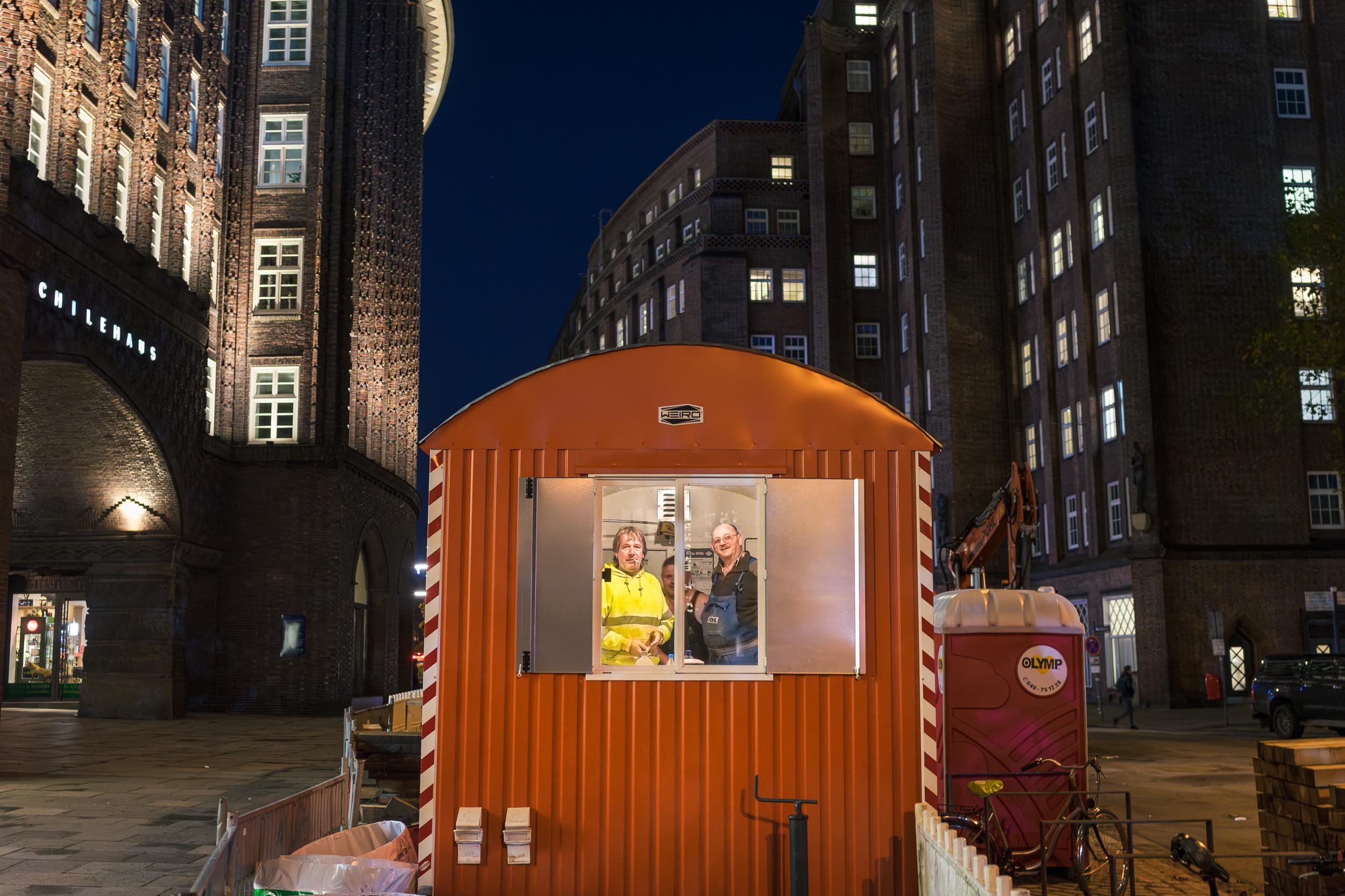 Two men smiling inside a small orange booth with open door and window, nighttime city street scene with large brick buildings, lit windows, and people walking in the background.
