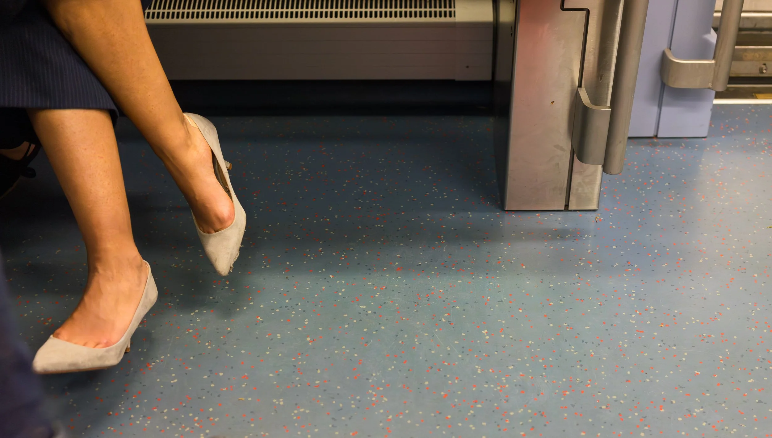 Close-up of a person sitting on a seat wearing beige high heels, with the floor showing a speckled pattern of small red, white, and yellow dots on a blue background.