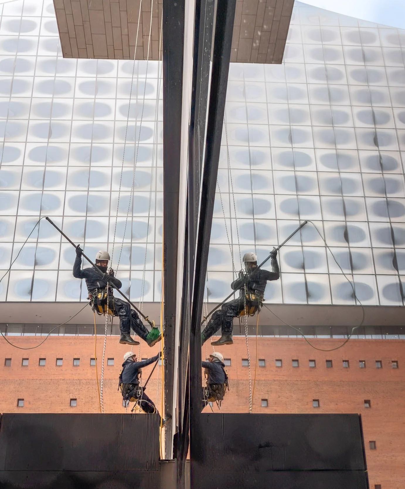 Two window cleaners wearing safety harnesses and helmets, cleaning a large glass building with a squeegee, with their reflection visible in the glass.