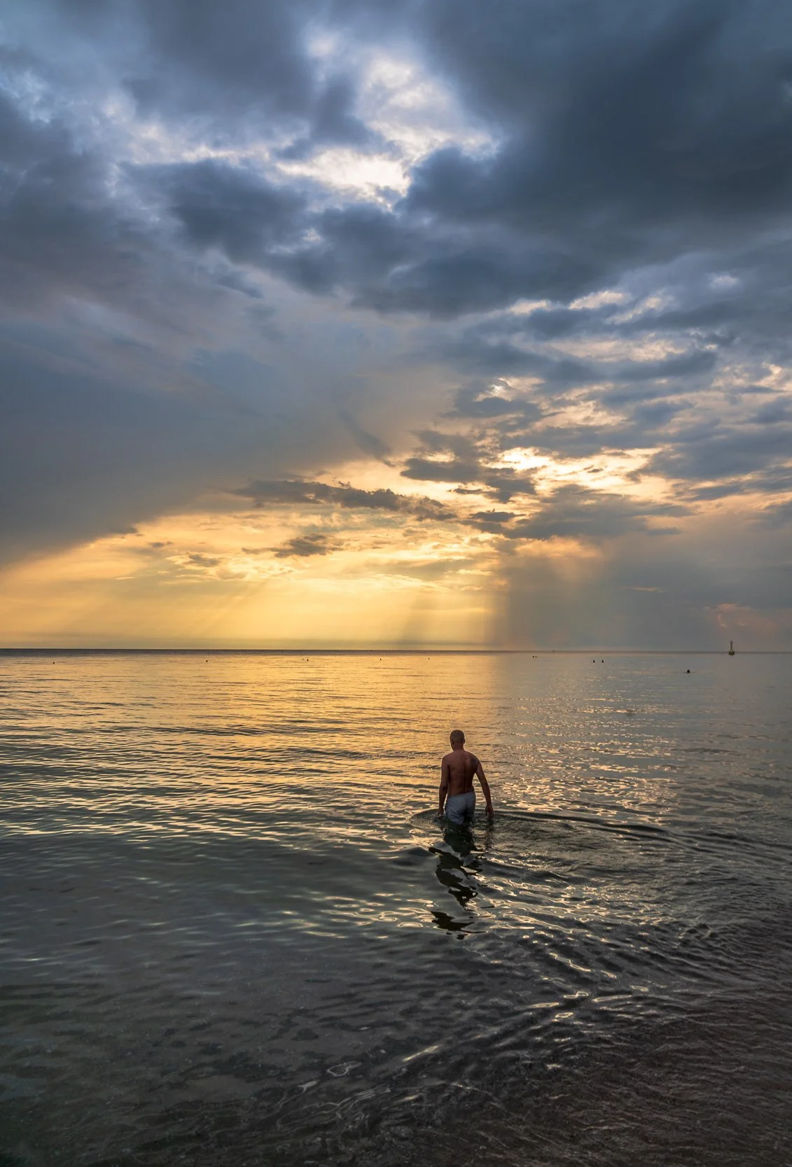 Man walking into the ocean at sunset with dramatic clouds in the sky.