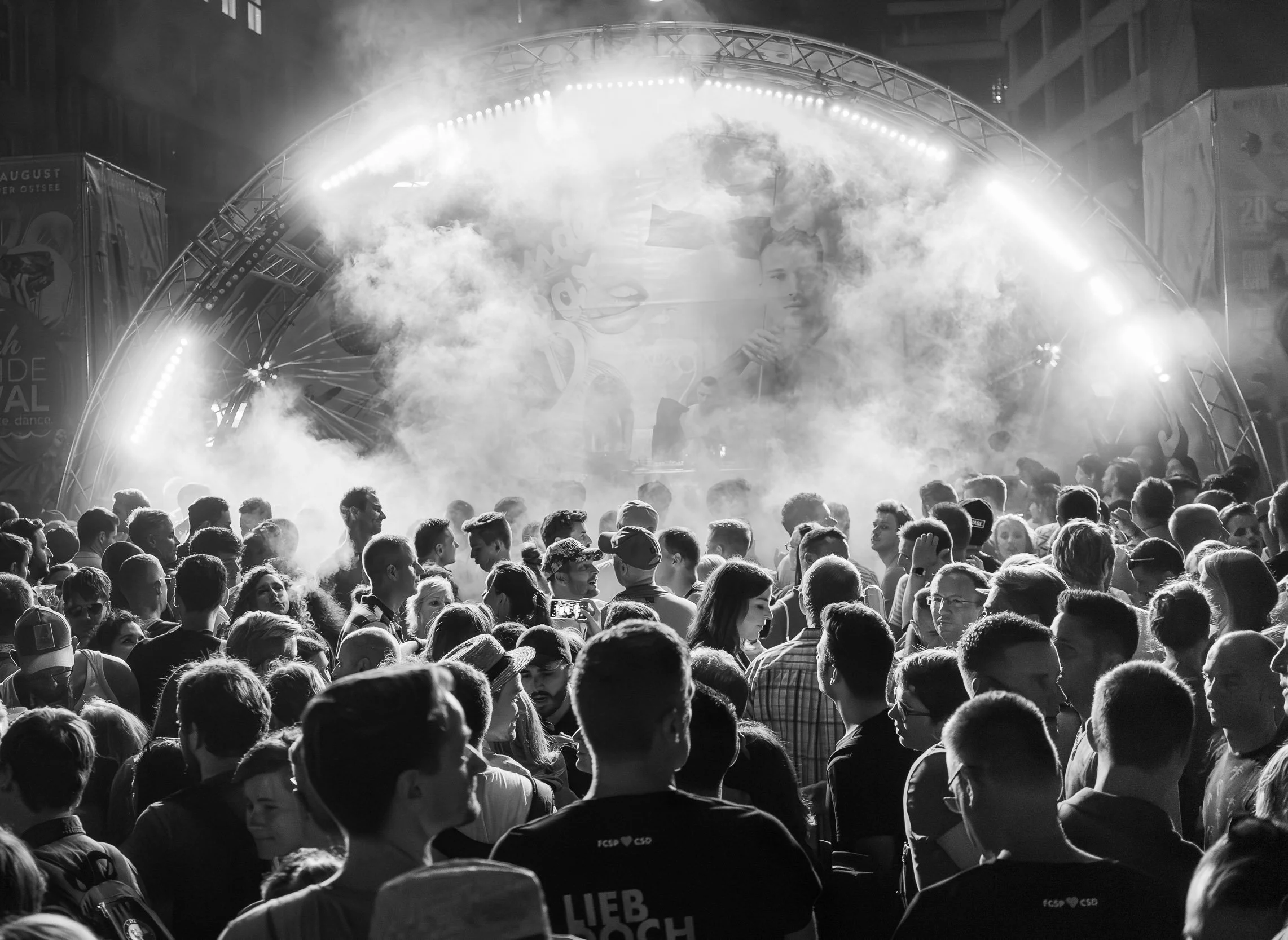 Crowd of people gathered at an outdoor concert or event at night, with a smoke machine and a stage with bright lights in the background.