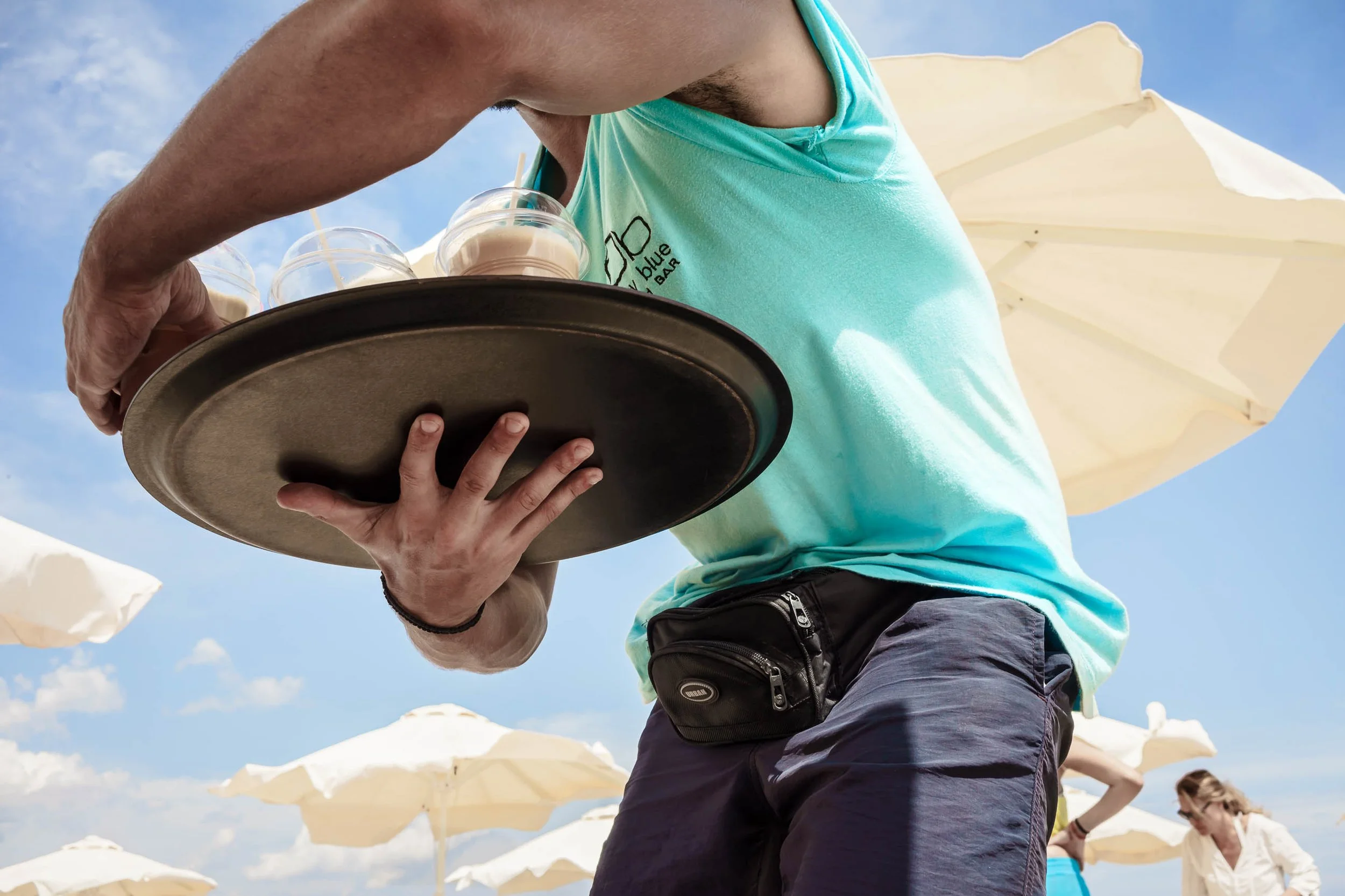 A person wearing a light blue sleeveless shirt with a logo, carrying a tray with drinks, holding it with their left hand, at a beach with umbrellas, under a partly cloudy sky.
