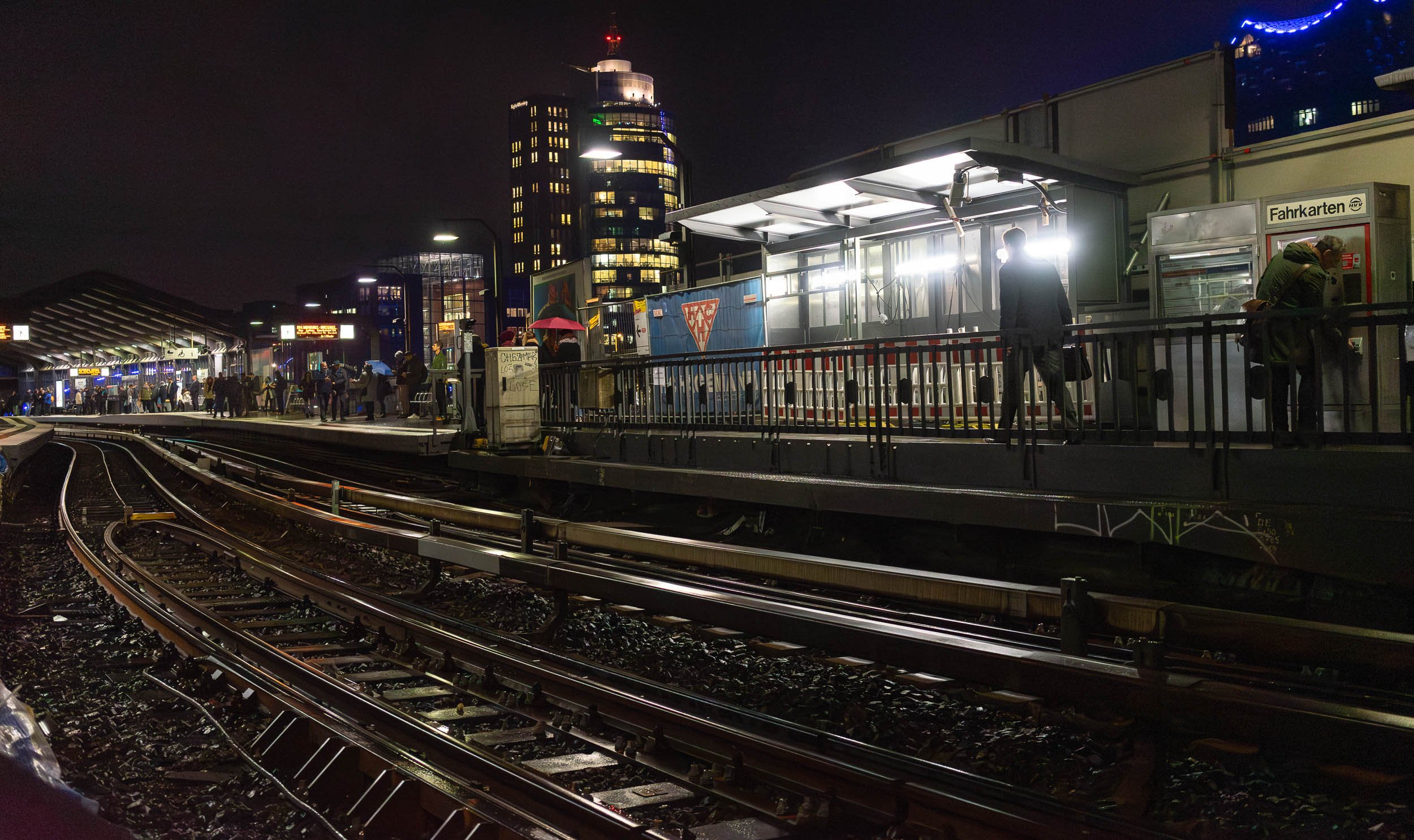 Night scene at a busy urban train station with people on the platform, illuminated buildings in the background, and train tracks in the foreground.